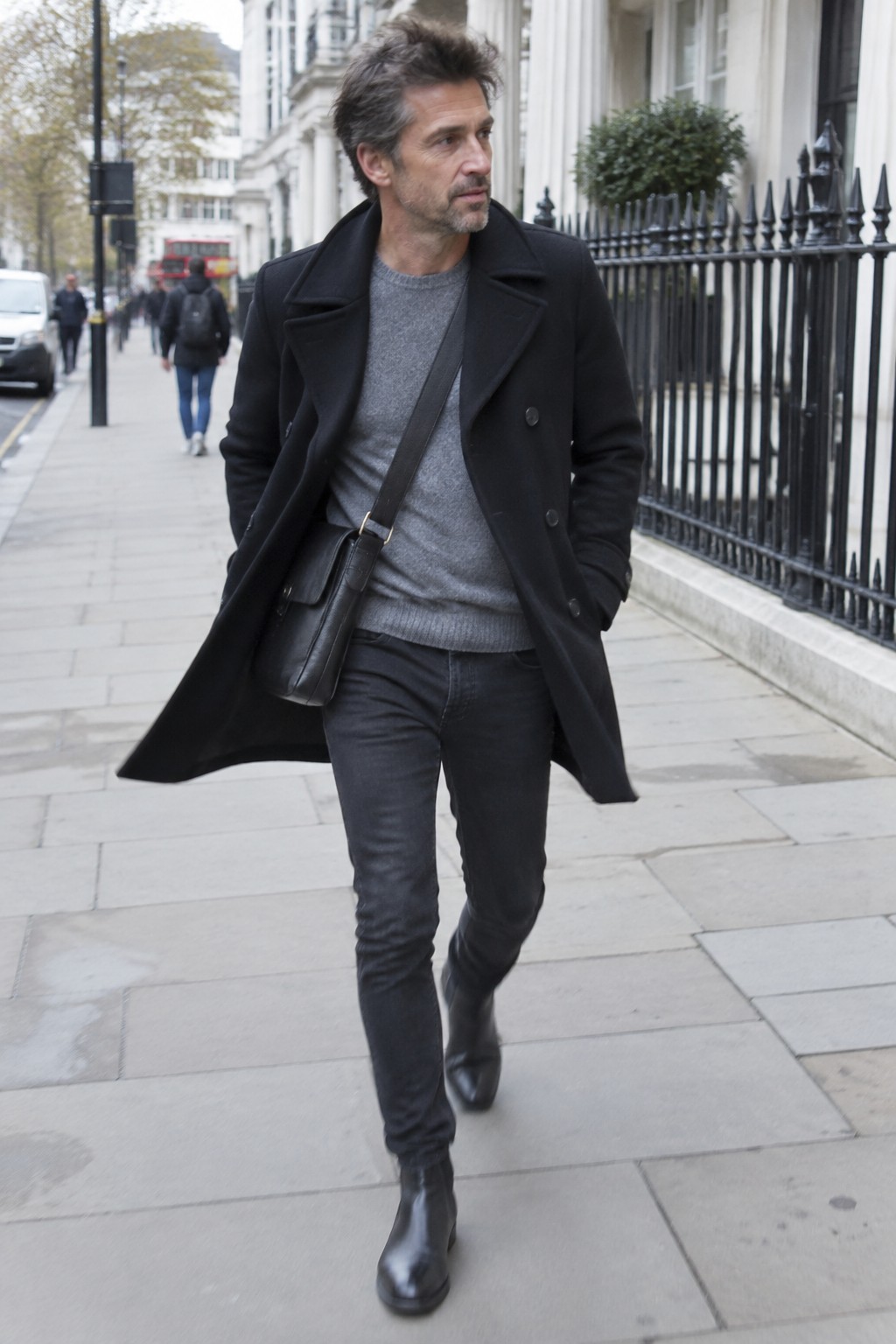 A man wearing a black double-breasted wool coat, gray crewneck sweater, dark charcoal jeans, black leather ankle boots, and a black crossbody bag.