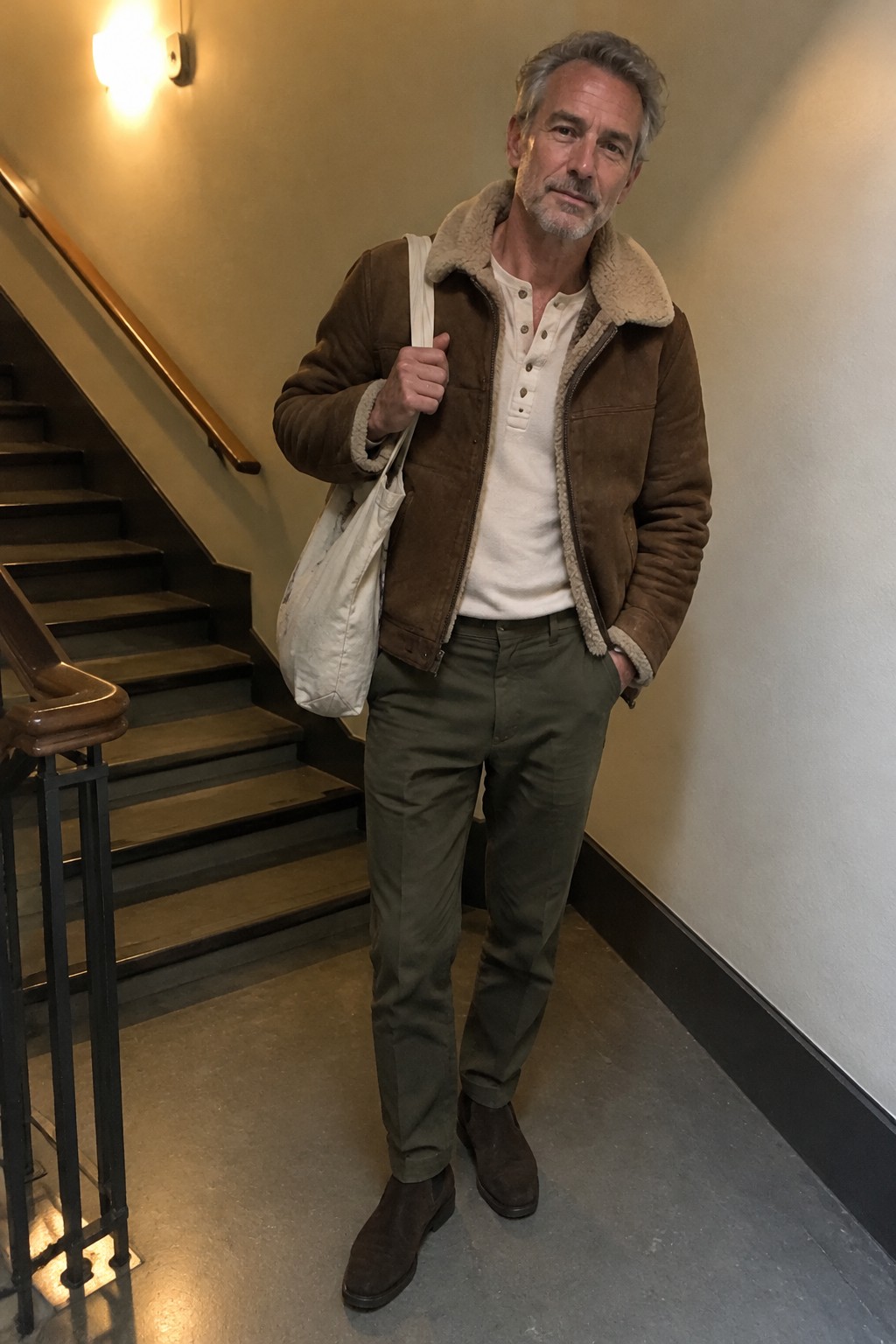 A gray-haired man wearing a brown shearling-trimmed suede jacket, cream henley shirt, olive trousers, dark brown shoes, and a white canvas shoulder bag.