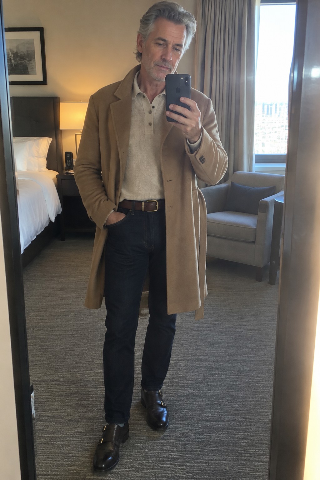 Man wearing a camel overcoat over a beige knit polo, dark jeans with turned-up hems, a brown belt, and black double-monk shoes.