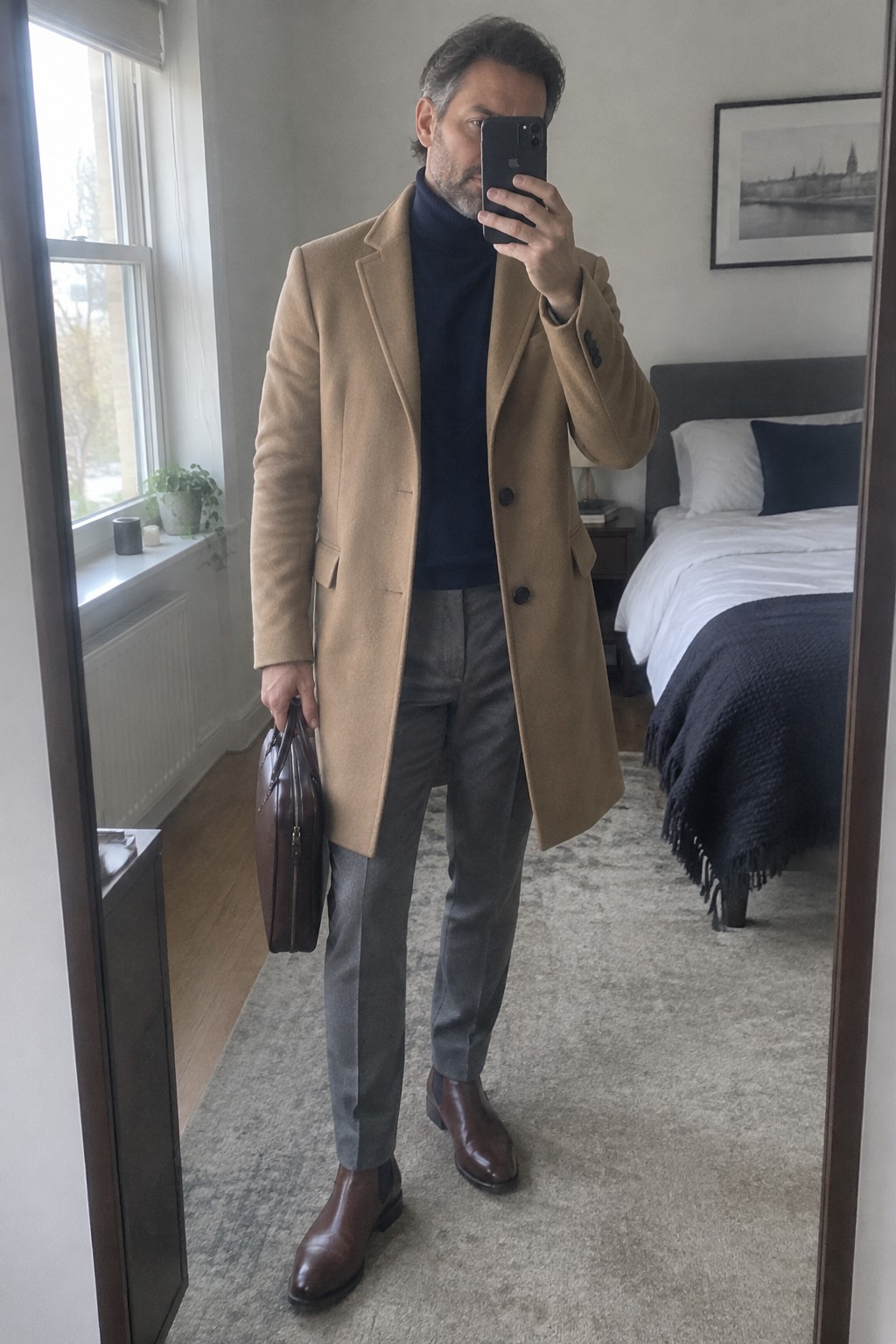 A man wears a camel overcoat over a navy turtleneck with gray trousers, brown leather Chelsea boots, and a dark brown briefcase.