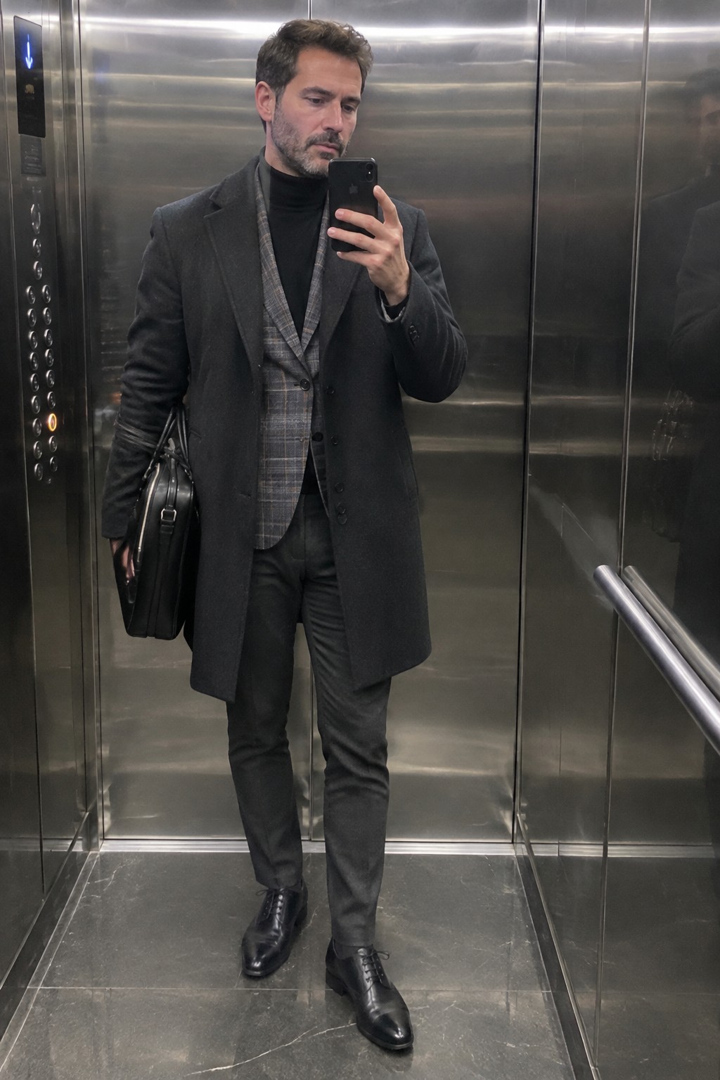 A man wears a charcoal overcoat over a black turtleneck and gray plaid waistcoat, with dark trousers, black leather lace-up shoes, and a black briefcase.