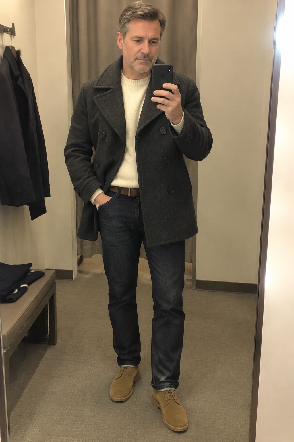 A man wearing a charcoal double-breasted peacoat over a cream ribbed sweater, dark jeans, a brown belt, and tan suede lace-up boots.