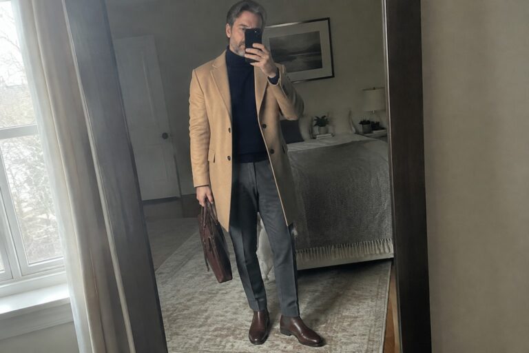 18 Confident Old Money Winter Outfits for Men Over 40 A man wears a camel overcoat over a navy turtleneck with gray trousers, brown leather Chelsea boots, and a dark brown briefcase.