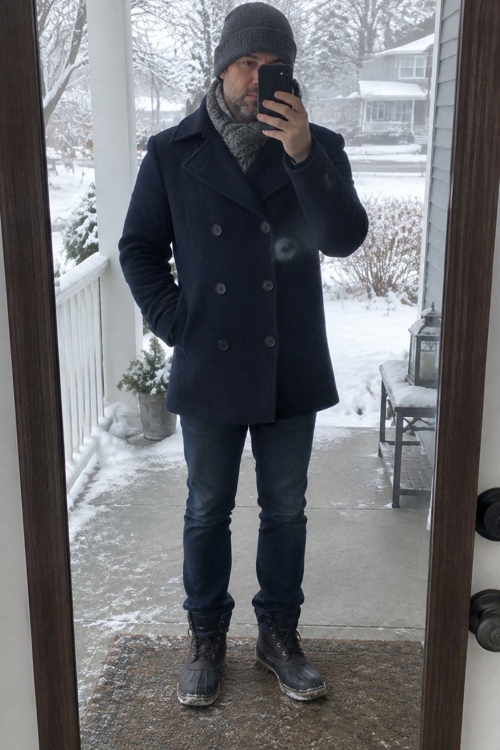 Man wearing a navy double-breasted peacoat over dark jeans, with a gray knit scarf, charcoal beanie, and dark lace-up winter boots.