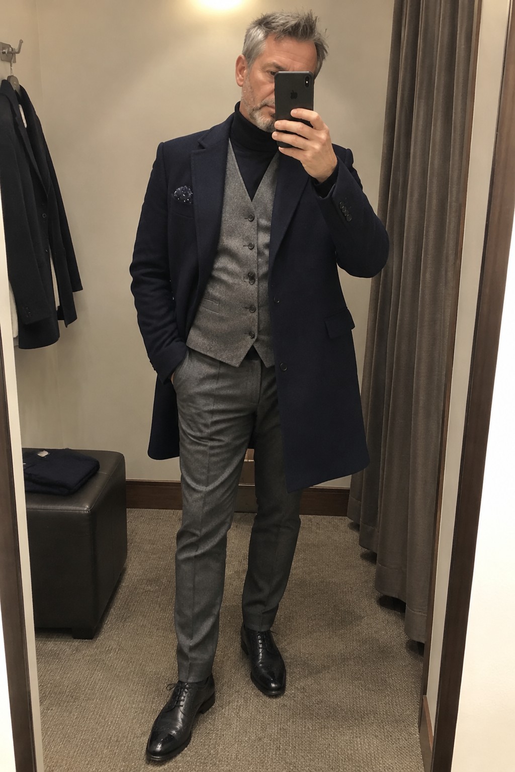 A man wearing a long navy topcoat over a black turtleneck, gray buttoned waistcoat, and gray trousers with black lace-up shoes.