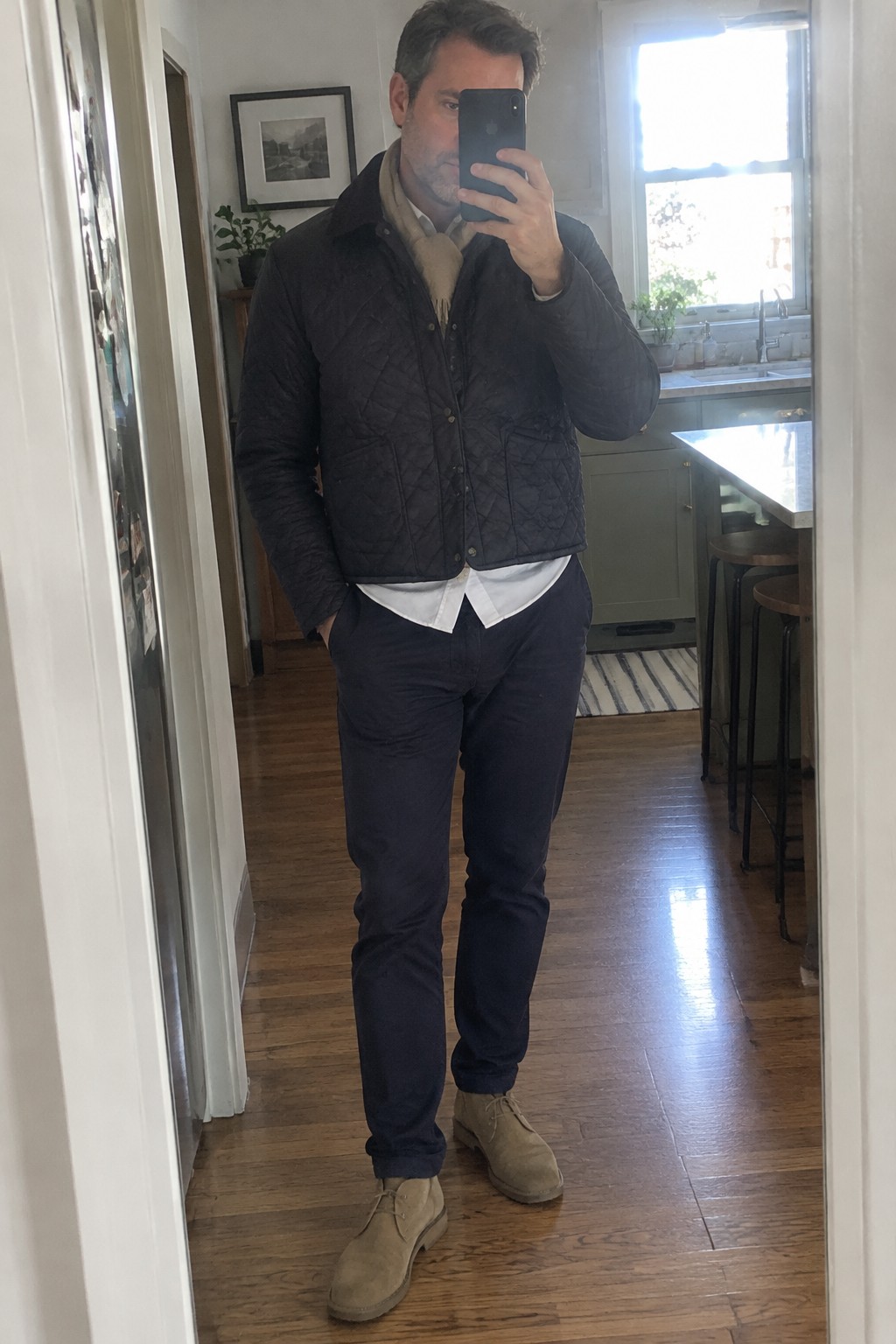Man wearing a dark quilted jacket over a white shirt and beige scarf, with navy trousers and tan suede chukka boots.