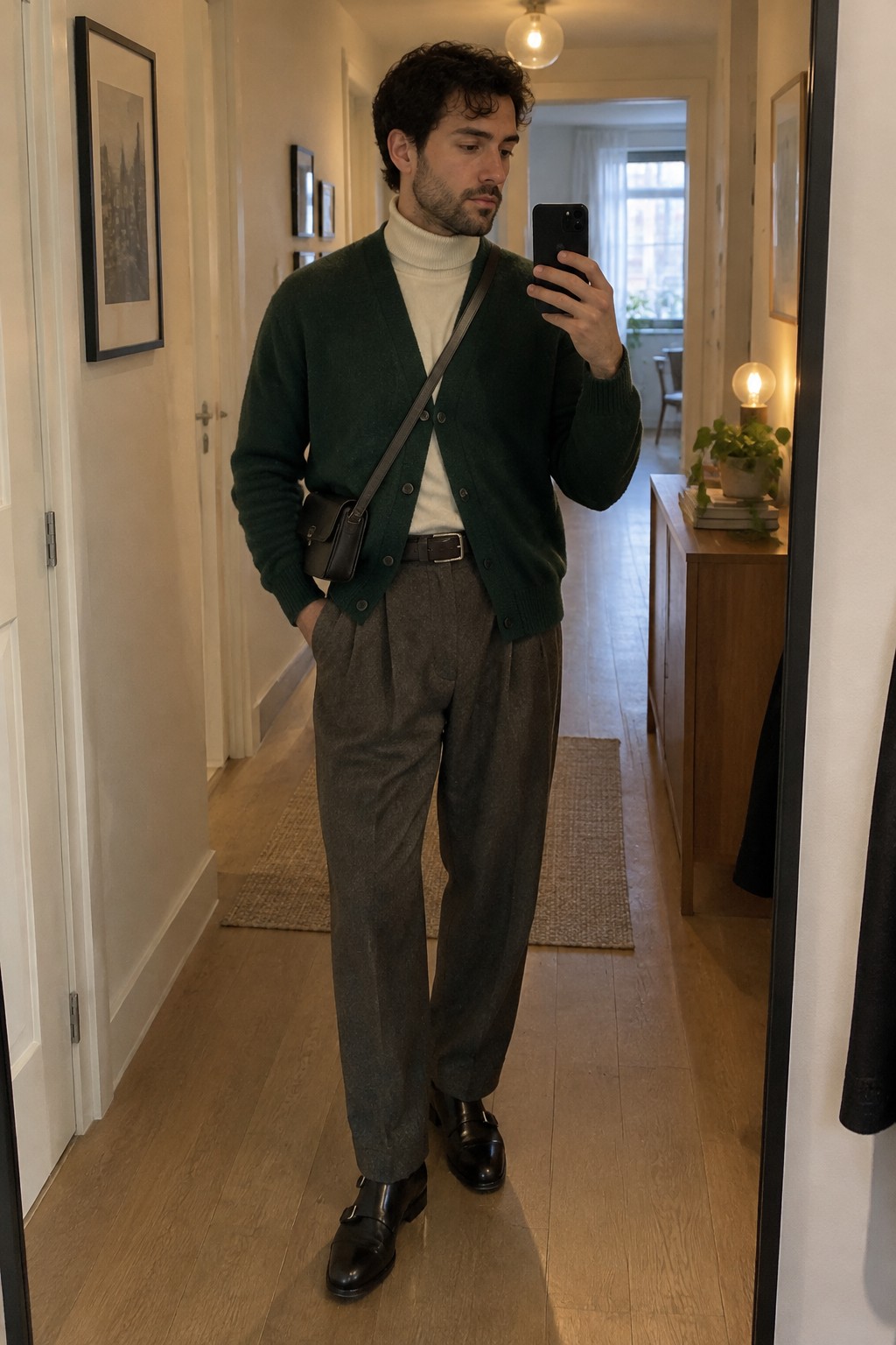 Man wearing a dark green cardigan over a cream turtleneck, gray pleated trousers, a black belt, a brown crossbody bag, and black buckle loafers.