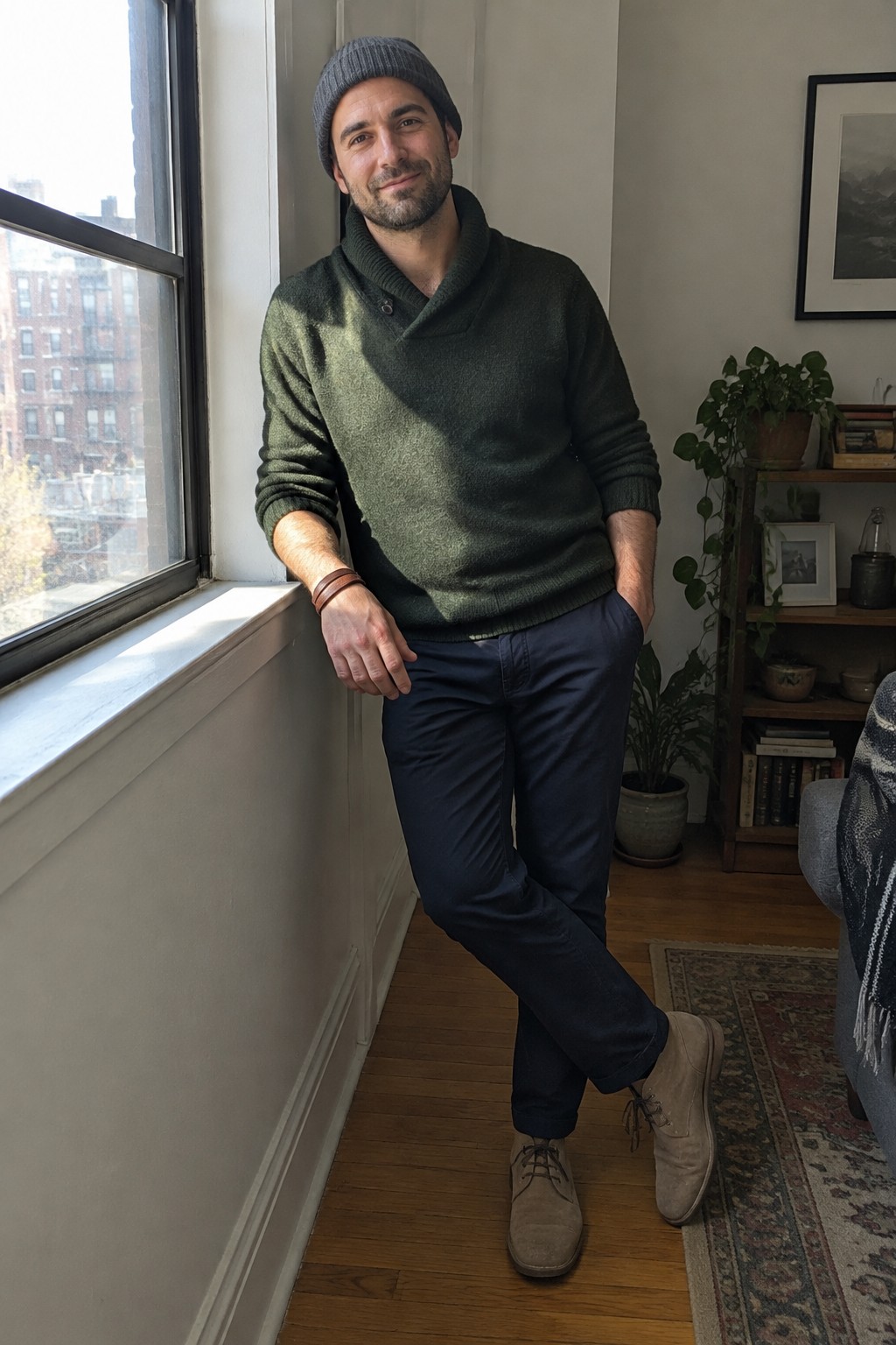 A man wearing a dark green shawl-collar sweater, navy trousers, a gray knit beanie, and taupe lace-up boots.