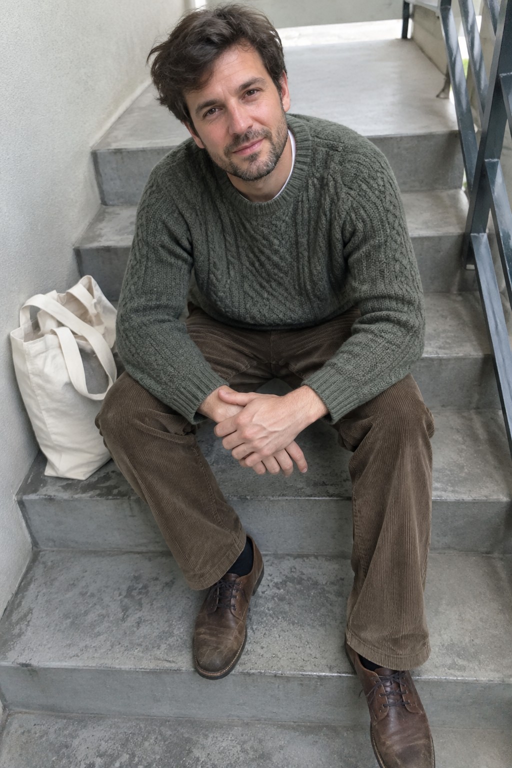Man seated on stone steps wearing a muted green cable-knit sweater over a white shirt, brown corduroy trousers, and brown leather lace-up shoes.
