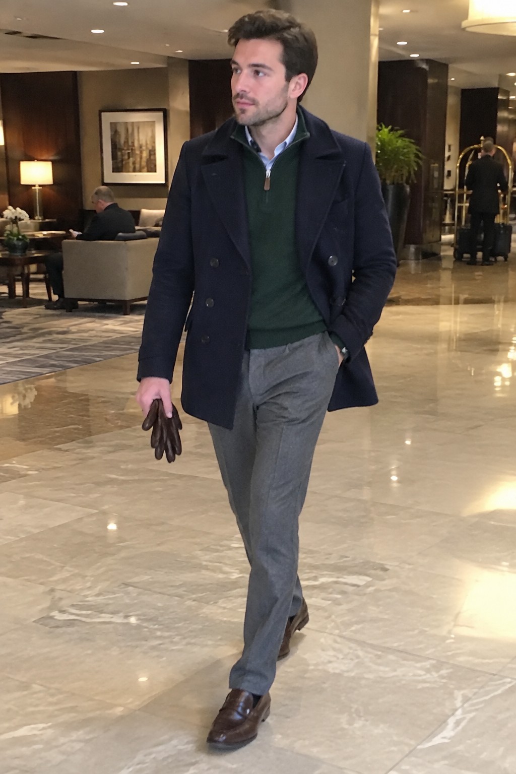 A man wearing a dark green quarter-zip sweater over a pale blue shirt, a navy double-breasted coat, gray trousers, brown leather loafers, and brown gloves.