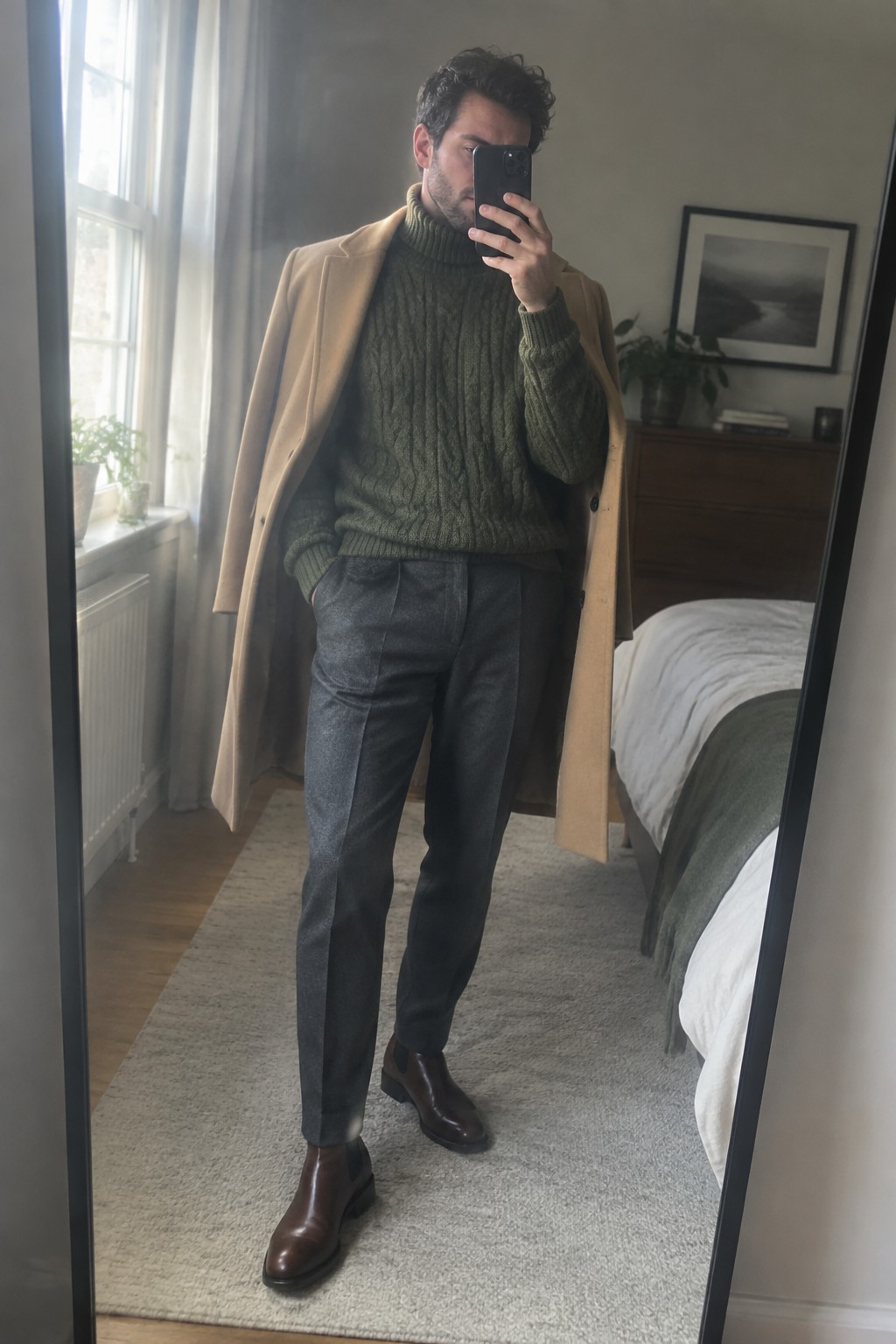 Man wearing an olive cable-knit turtleneck, camel overcoat, charcoal trousers, and dark brown Chelsea boots.