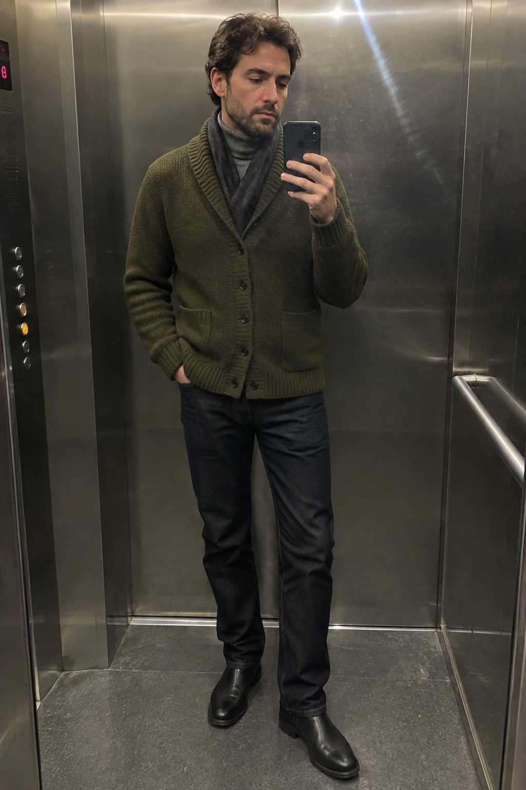 A man wearing an olive green shawl-collar cardigan over a gray turtleneck and dark scarf, with dark jeans and black leather boots.