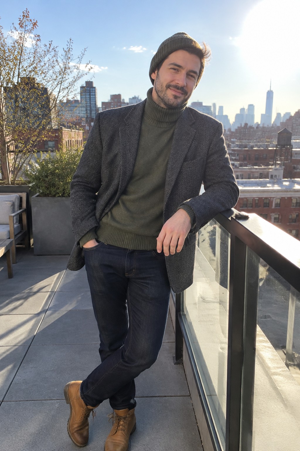 A man wearing an olive green ribbed turtleneck, charcoal herringbone overcoat, dark cuffed jeans, brown lace-up boots, and a dark knit beanie.