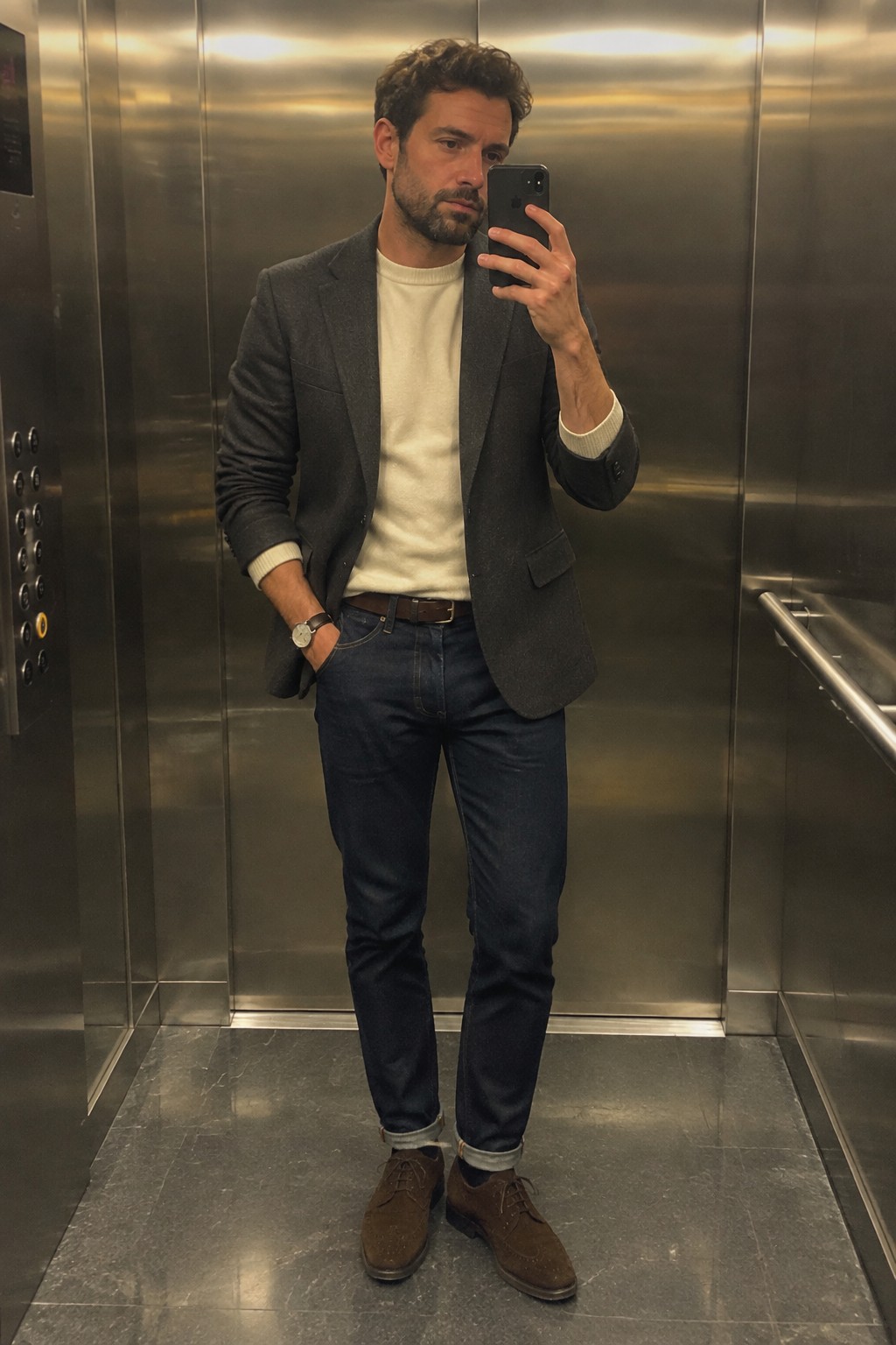 A man wears a charcoal blazer over a cream sweater with dark cuffed jeans, a brown belt, brown suede lace-up shoes, and a wristwatch.