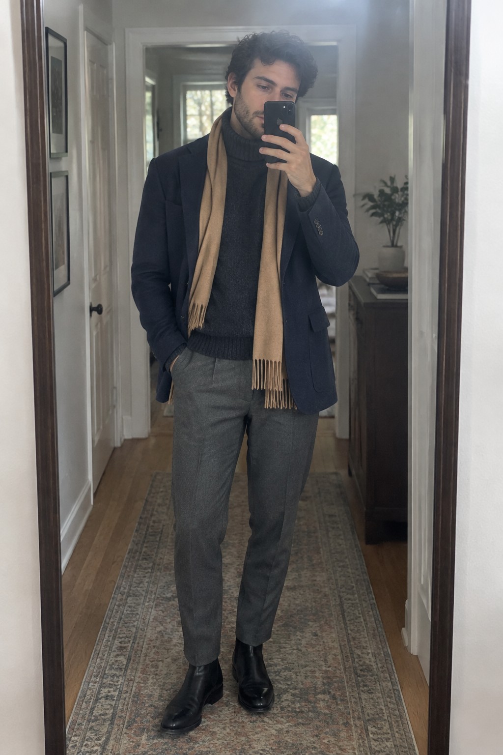 Man wearing a navy blazer over a charcoal turtleneck, a camel scarf, gray trousers, and black leather ankle boots.