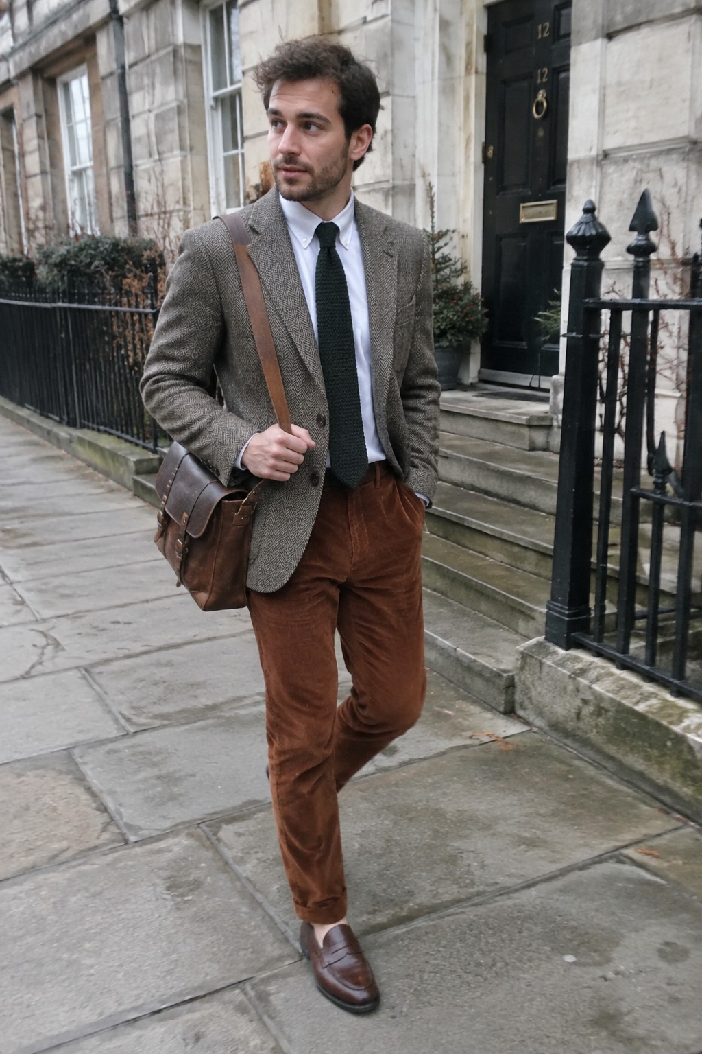 A man wears a gray herringbone blazer, white shirt, dark green tie, rust-brown corduroy trousers, brown leather loafers, and a brown leather crossbody bag.