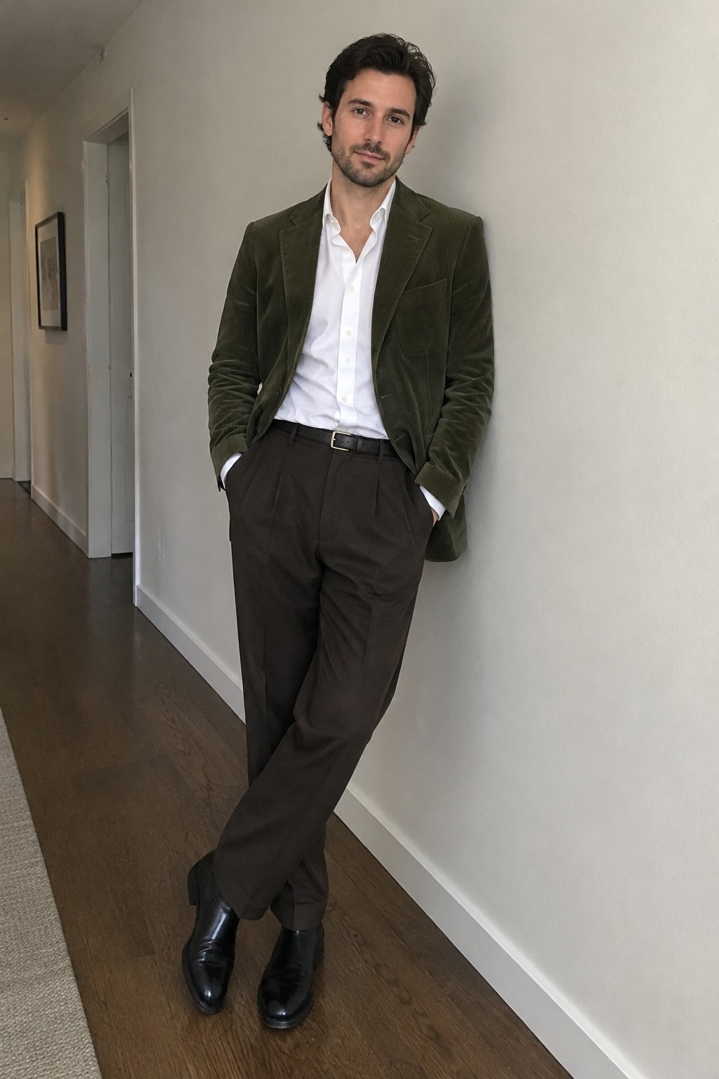 A man wearing a moss green corduroy blazer over a white button-down shirt, brown pleated trousers with a black belt, and black ankle boots.