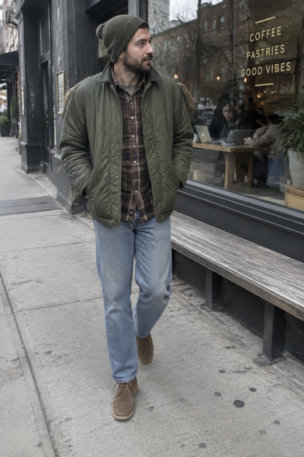A man wearing a moss green quilted jacket, brown plaid shirt, charcoal knit beanie, light-wash jeans, and brown suede boots.