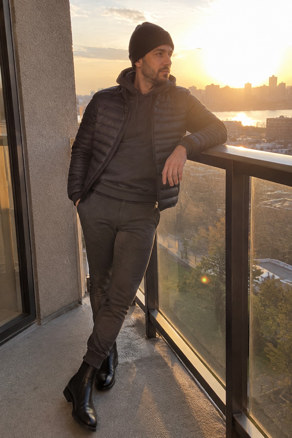 A man wearing a black beanie, black quilted puffer jacket over a dark hoodie, slim charcoal trousers, and black ankle boots.