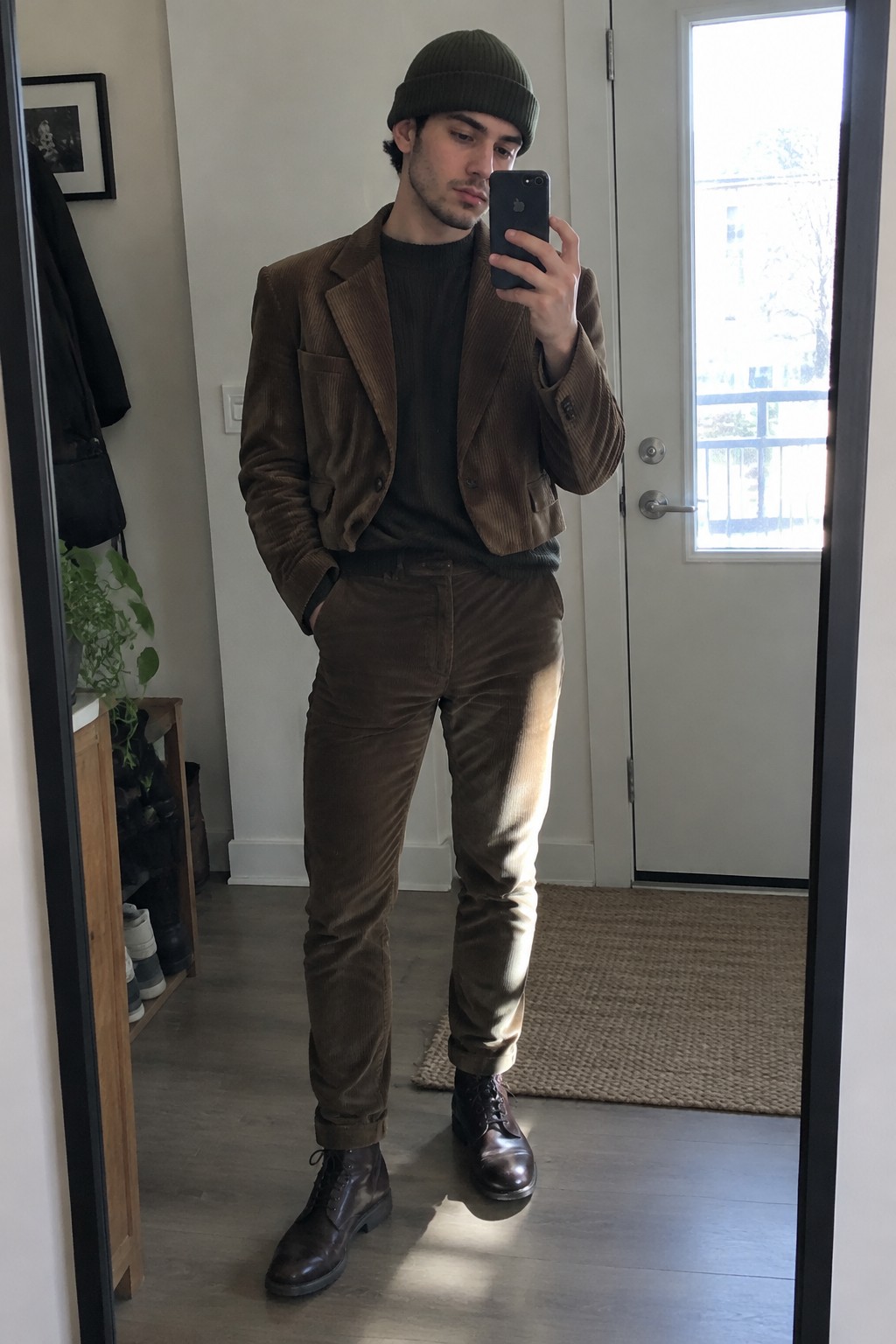 A man wearing a cropped brown corduroy jacket and matching trousers with a dark olive sweater, ribbed beanie, and dark lace-up boots.