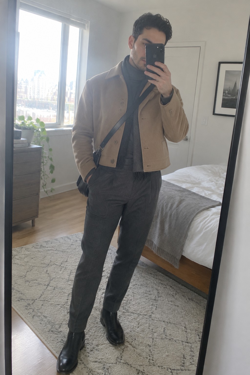 A man wearing a cropped camel jacket over a charcoal turtleneck and tapered charcoal trousers, with black leather ankle boots and a black crossbody bag.