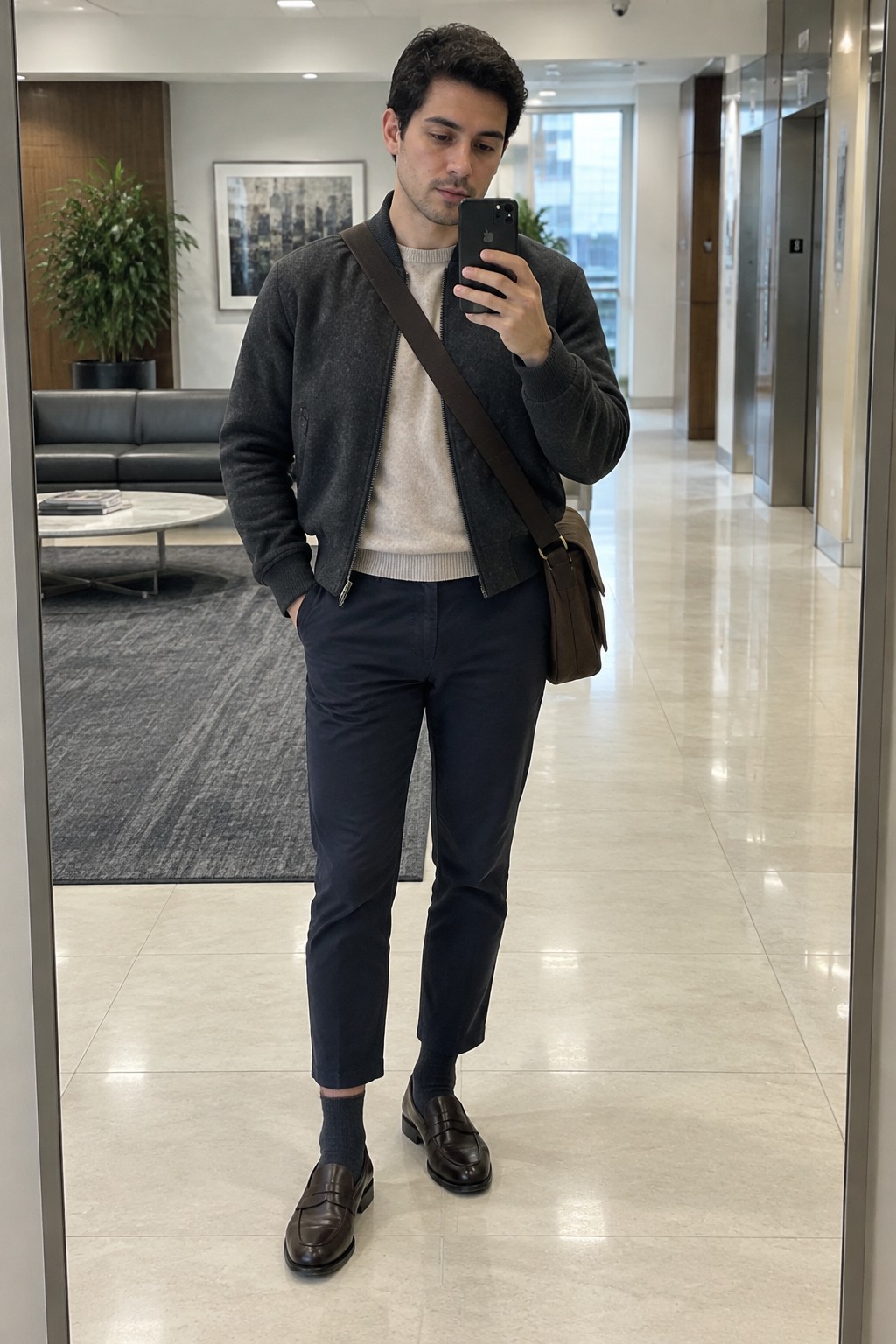 A man wearing a charcoal bomber jacket over a cream sweater, cropped navy trousers, dark socks, brown penny loafers, and a brown crossbody bag.