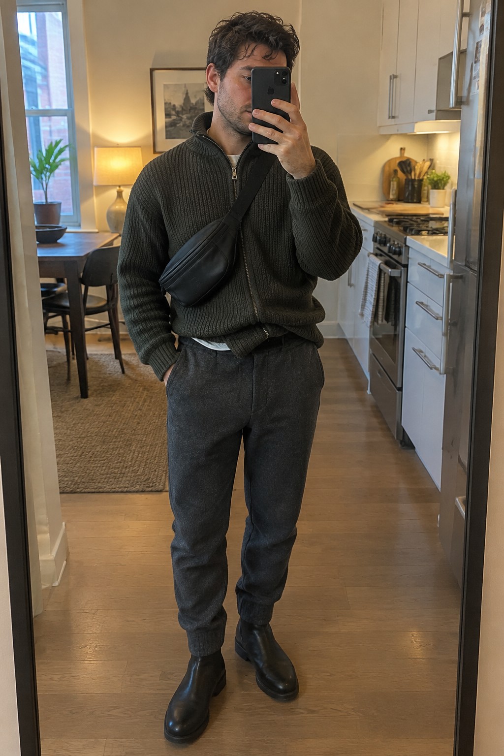 A man taking a mirror selfie in a charcoal ribbed zip-up sweater, dark tapered trousers, black Chelsea boots, and a black crossbody bag.