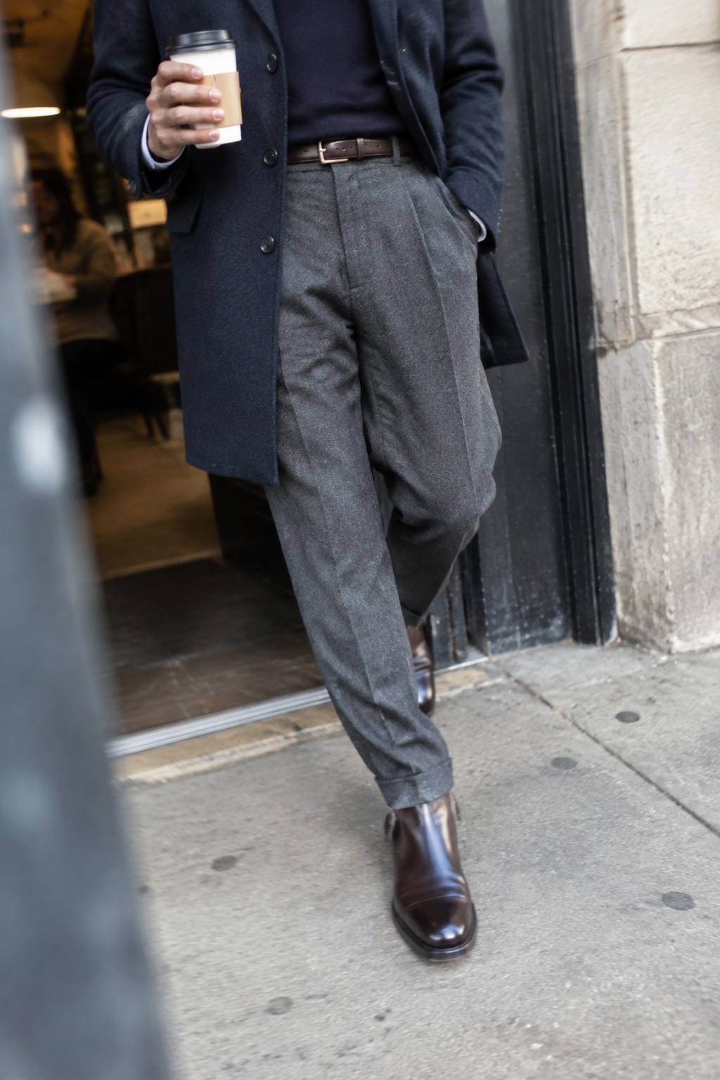 A person wearing a navy overcoat over a dark sweater, high-waisted pleated gray trousers, a brown belt, and polished brown ankle boots, holding a takeaway coffee cup.