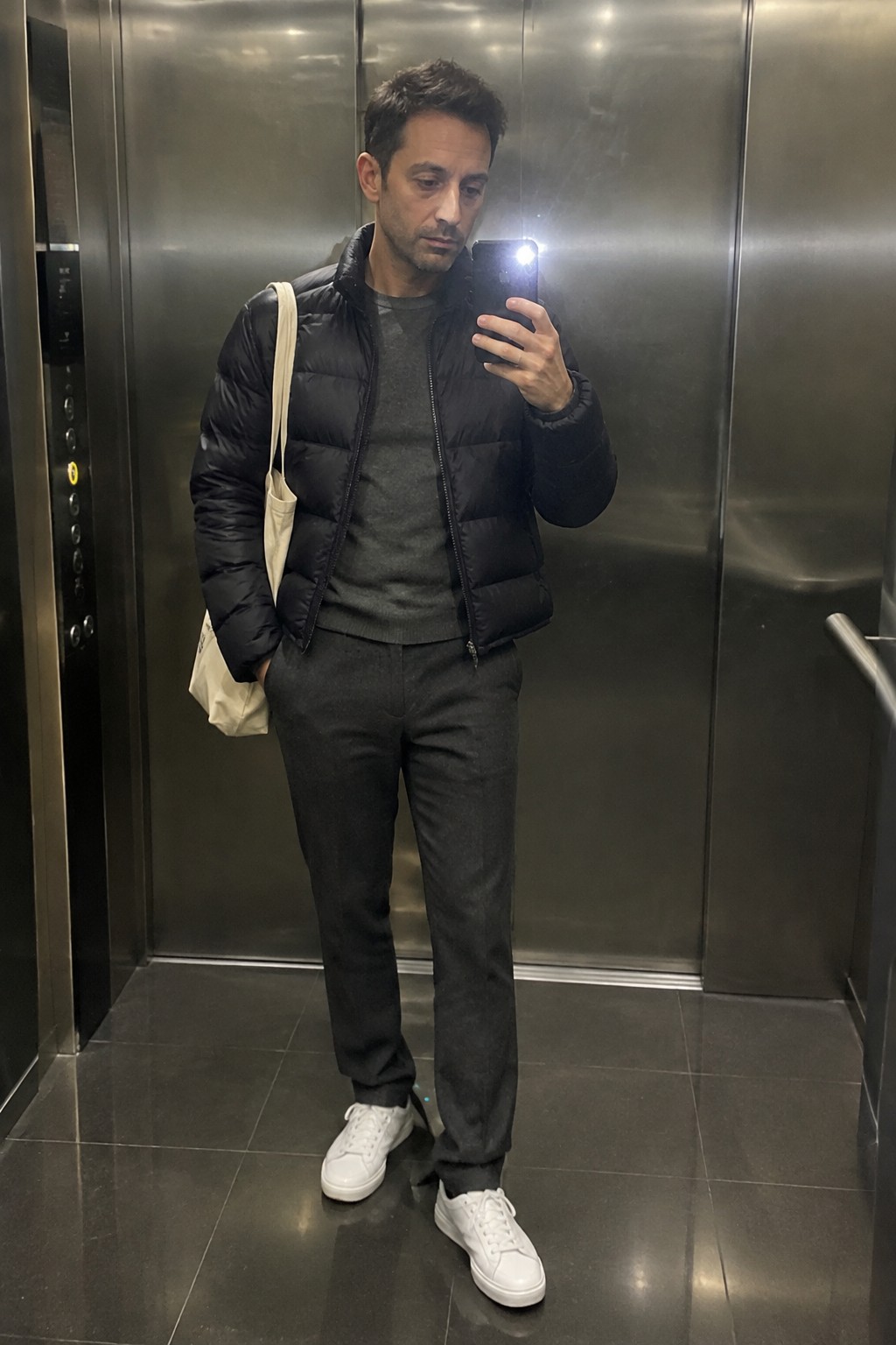 A man wears a black quilted puffer jacket over a charcoal sweater with dark gray tailored trousers, white sneakers, and a cream canvas tote.