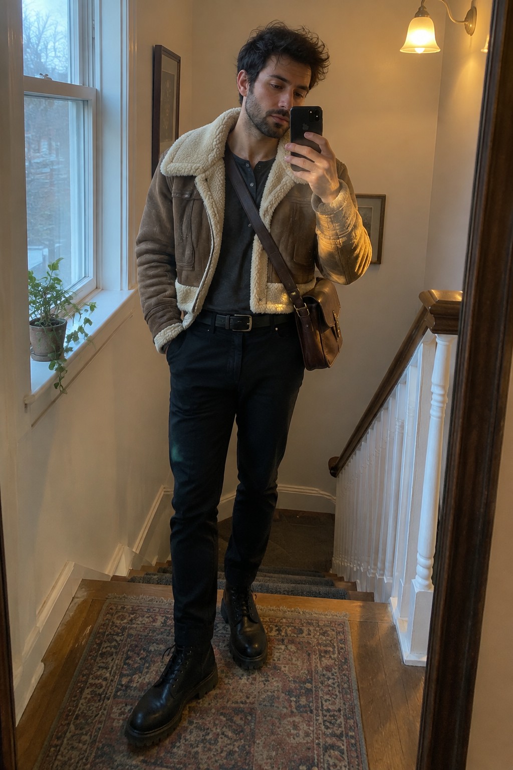 A man wearing a brown shearling-lined jacket, black henley, slim black jeans, black lace-up boots, and a brown leather crossbody bag.