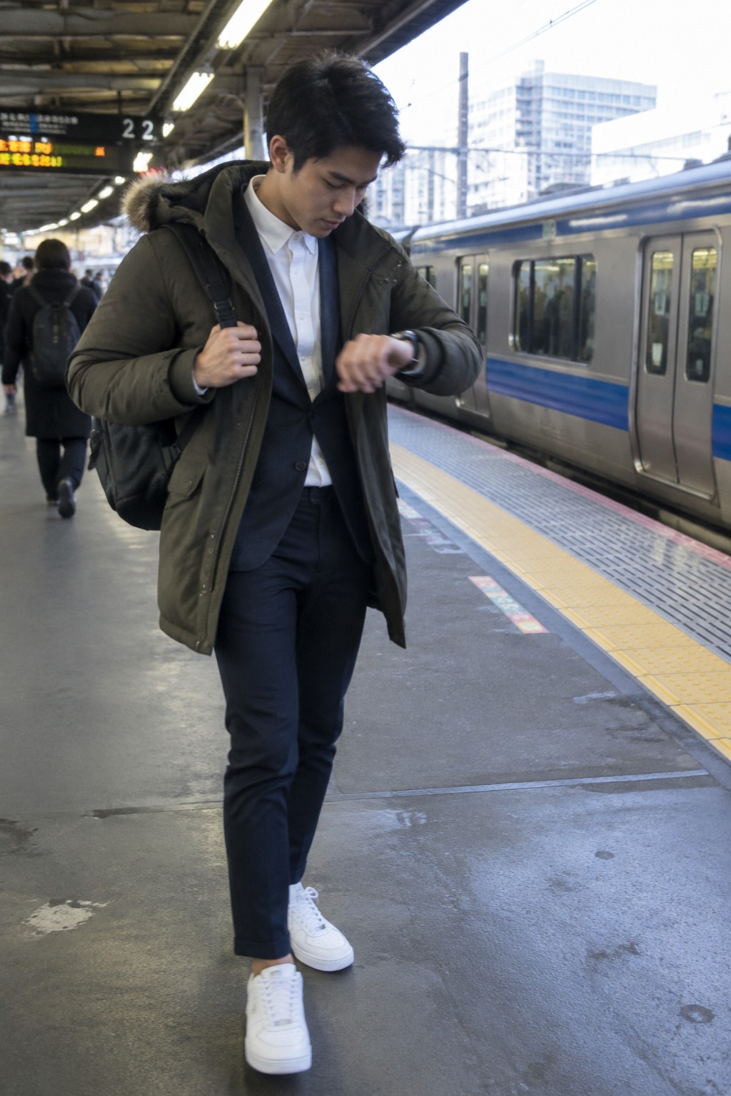 A man in an olive fur-trimmed parka over a navy suit and white shirt, wearing cropped navy trousers, white sneakers, and a black backpack.