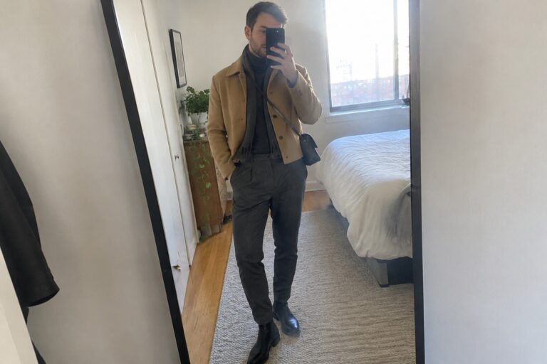 A man wearing a cropped camel jacket over a charcoal turtleneck and tapered charcoal trousers, with black leather ankle boots and a black crossbody bag.
