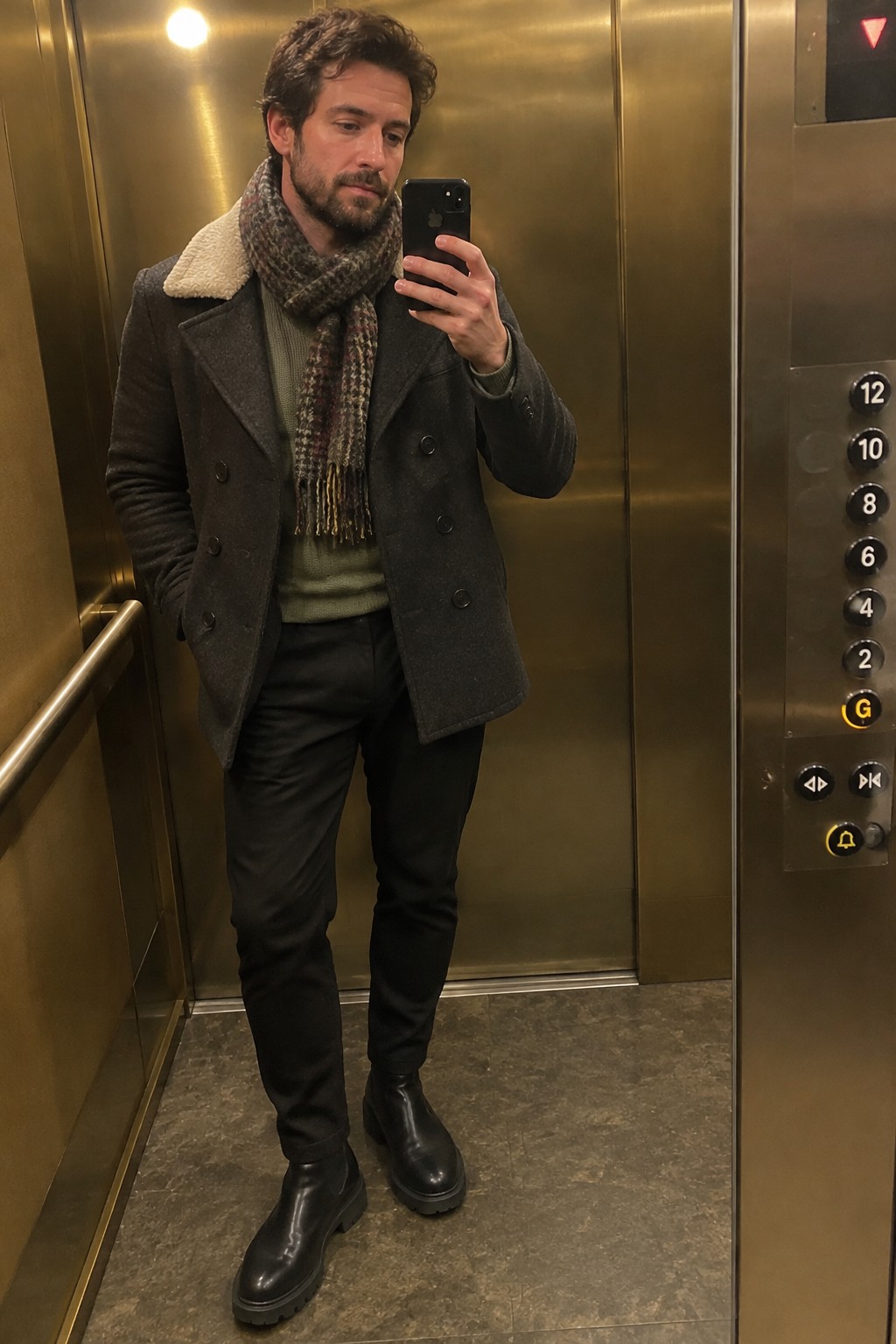 Man wearing a charcoal double-breasted wool coat with a shearling collar, patterned scarf, olive sweater, black trousers, and black Chelsea boots.
