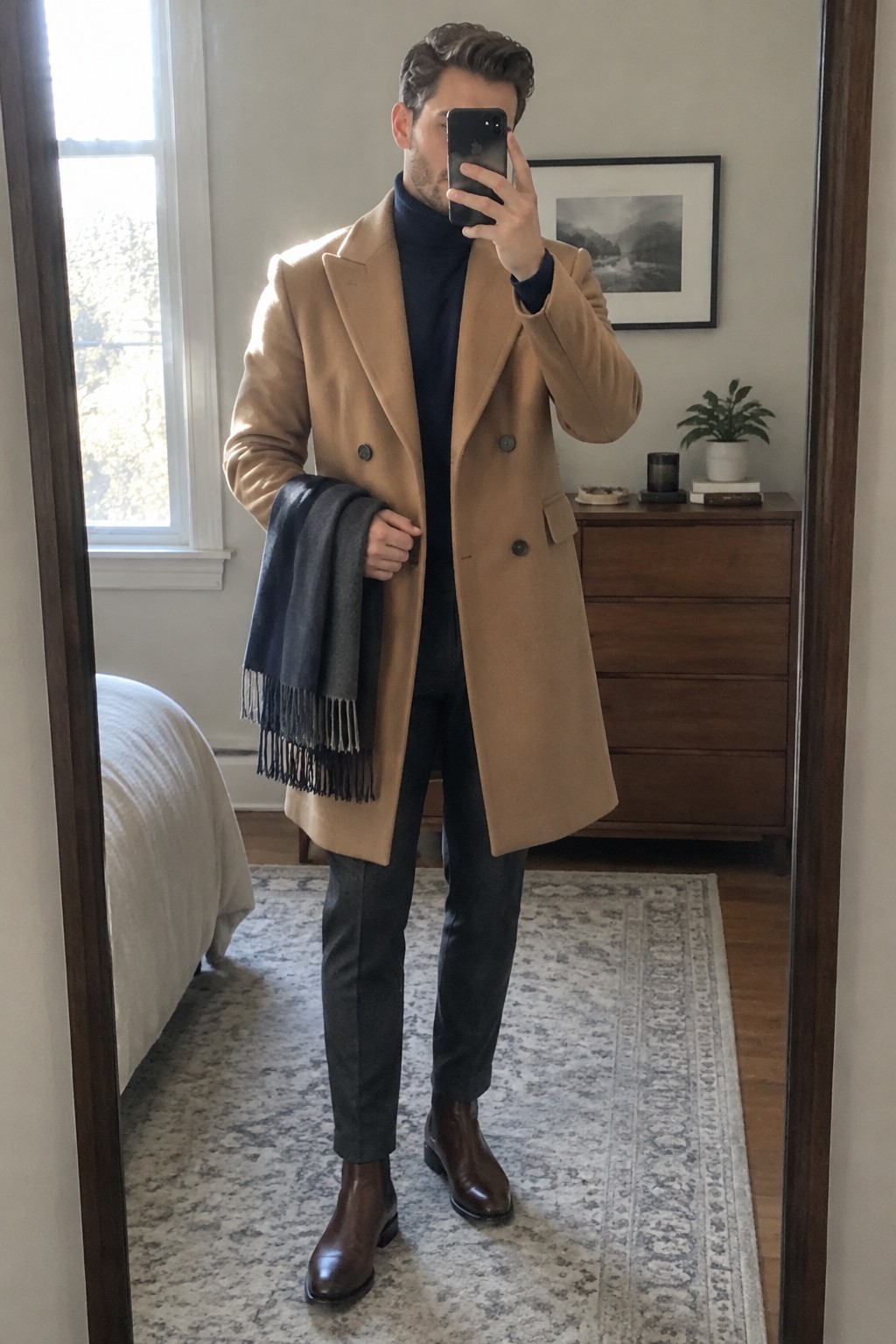 A man wears a camel double-breasted overcoat over a navy turtleneck and charcoal trousers, with brown Chelsea boots and a gray scarf.