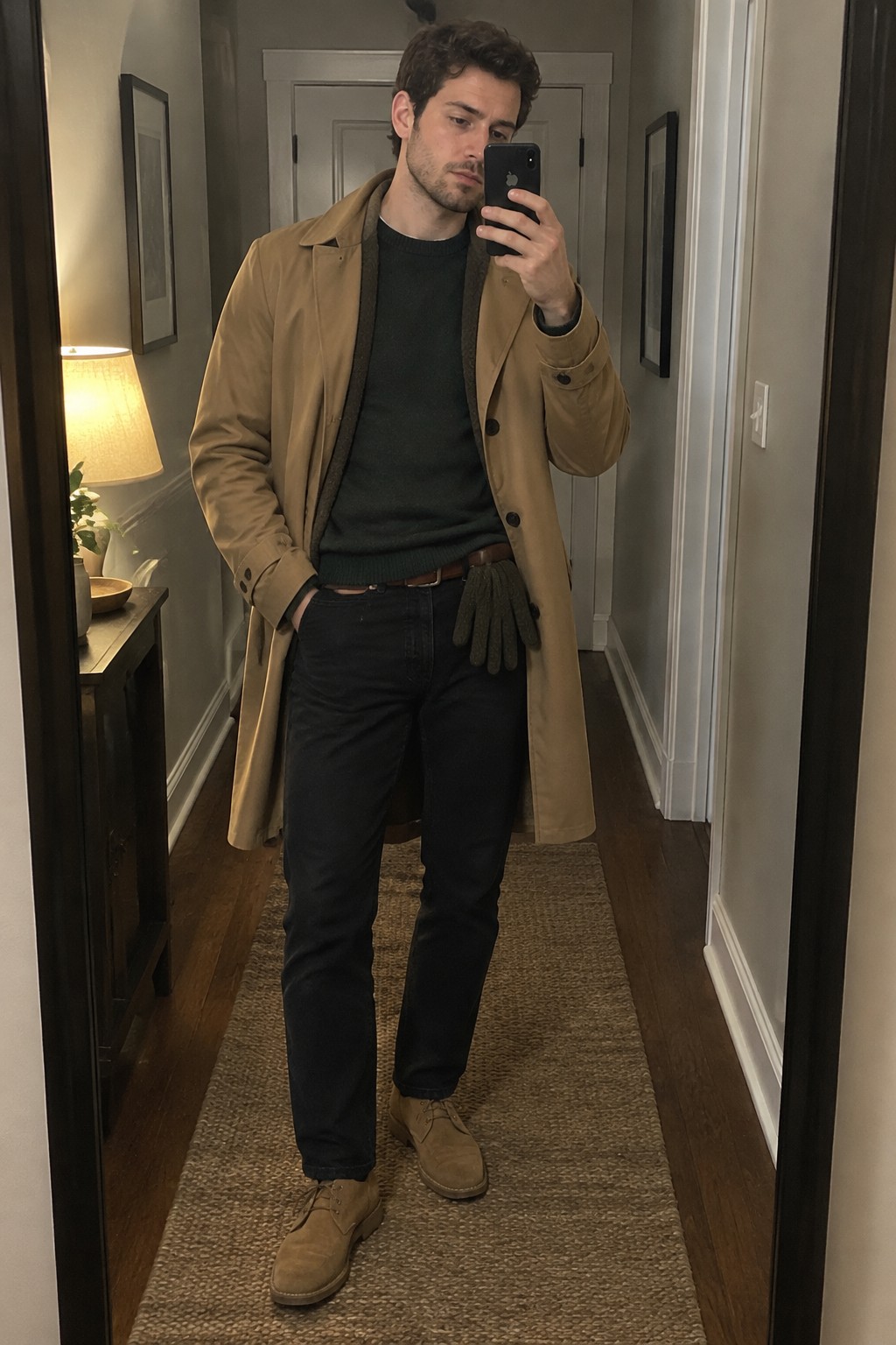A man wearing a camel trench coat over a charcoal sweater and dark jeans, with brown suede lace-up boots and gloves tucked at his waistband.