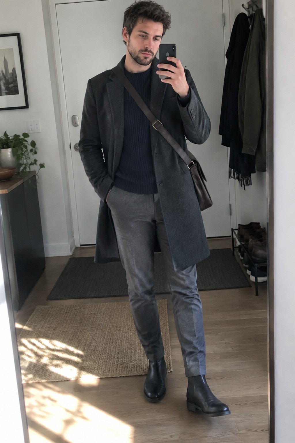 A man wears a charcoal overcoat over a navy ribbed sweater, grey trousers, black Chelsea boots, and a brown crossbody bag.