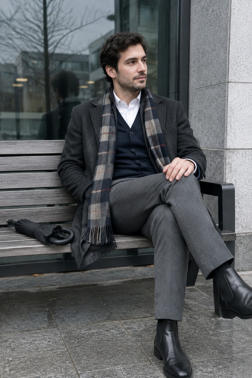 Man wearing a charcoal wool coat, navy waistcoat, white shirt, gray trousers, plaid scarf, black Chelsea boots, and black gloves.