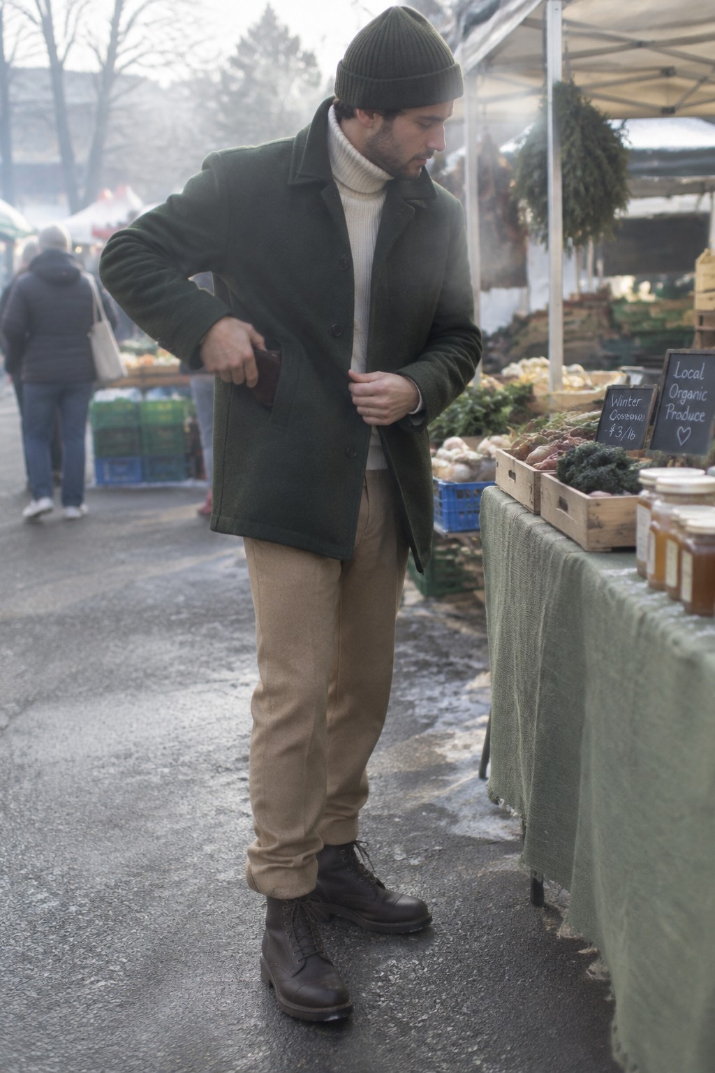 A man wearing a dark green wool coat, cream ribbed turtleneck, taupe trousers, dark knit beanie, and black lace-up boots.