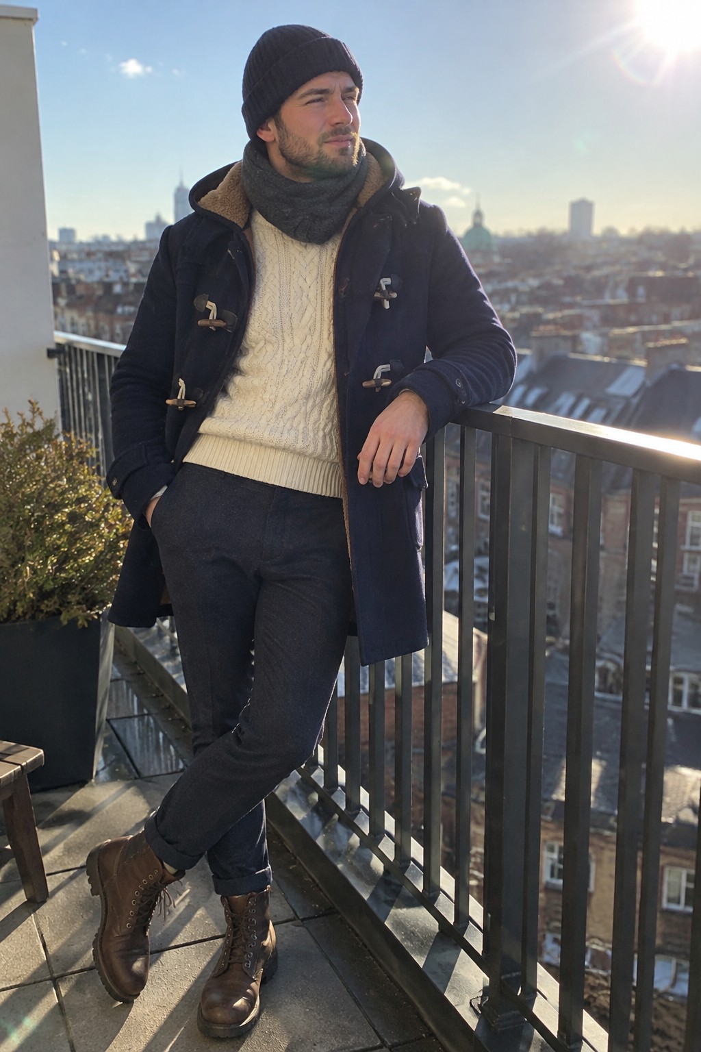 A man wearing a navy duffle coat over a cream cable-knit sweater, with a charcoal scarf, black beanie, dark cuffed trousers, and brown lace-up boots.