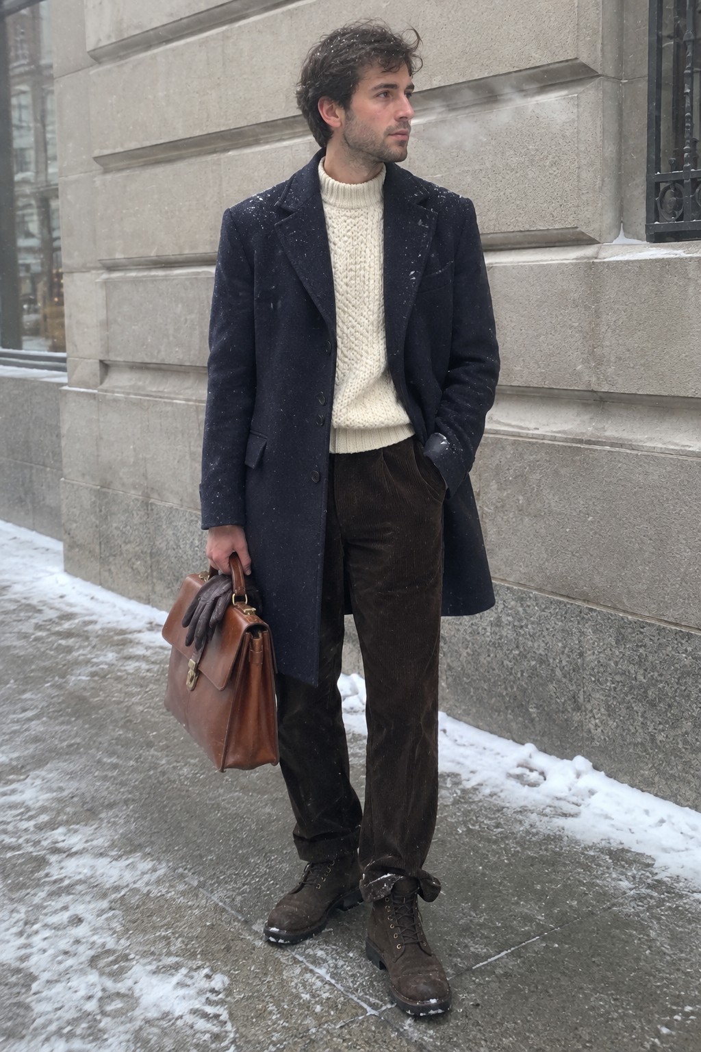 A man in a navy wool coat, cream cable-knit sweater, brown corduroy trousers, and brown lace-up boots carries a leather briefcase and gloves.