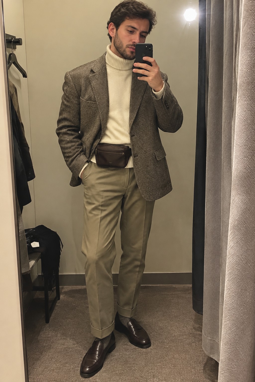 Man wearing a brown tweed blazer over a cream turtleneck, olive-beige cuffed trousers, a dark brown belt bag, and brown loafers.