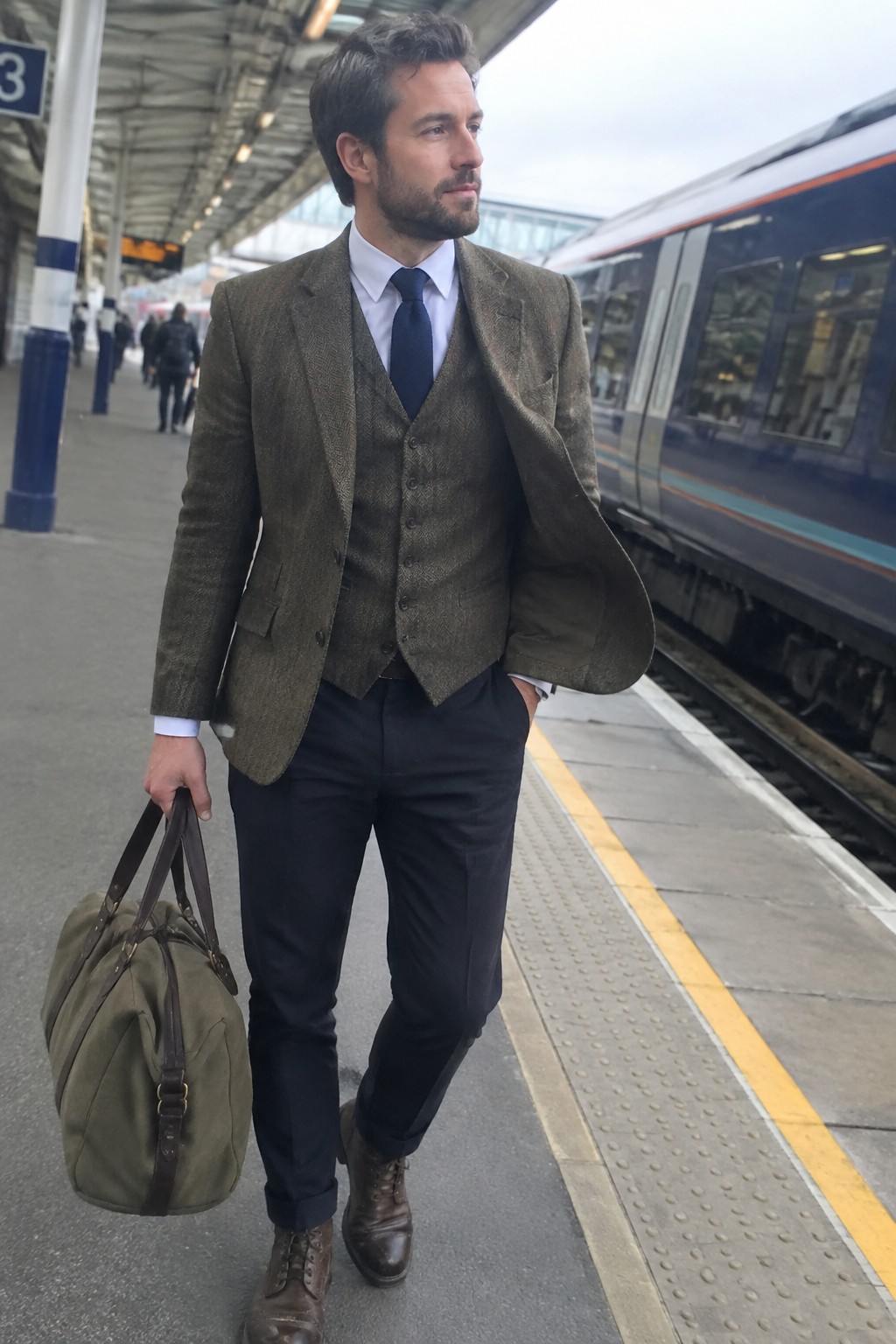 A man wearing an olive-brown tweed blazer and matching waistcoat with a white shirt, navy tie and trousers, brown lace-up boots, and an olive duffel bag.