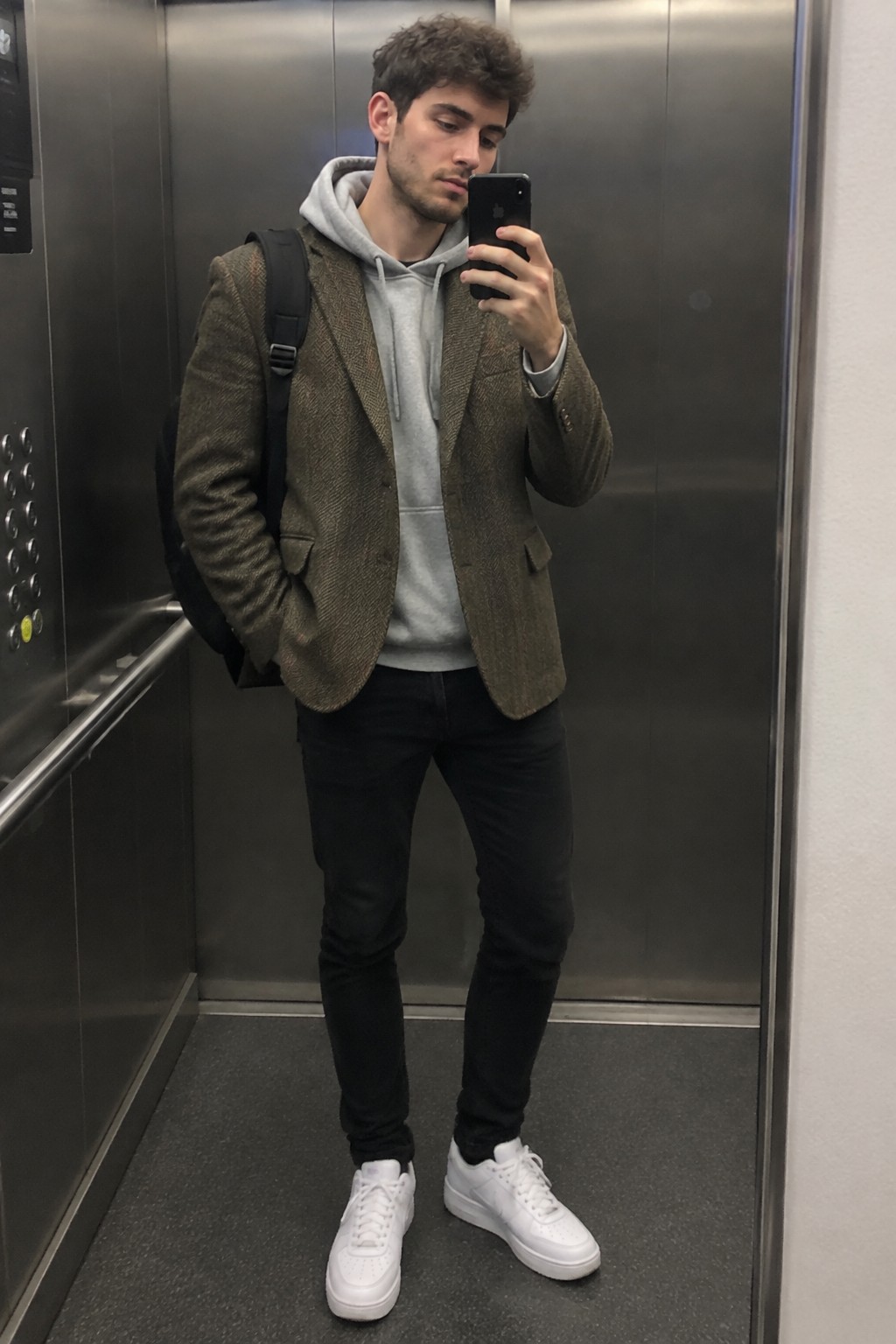 A man wearing an olive-brown tweed blazer over a grey hoodie, black jeans, white trainers, and a black backpack.