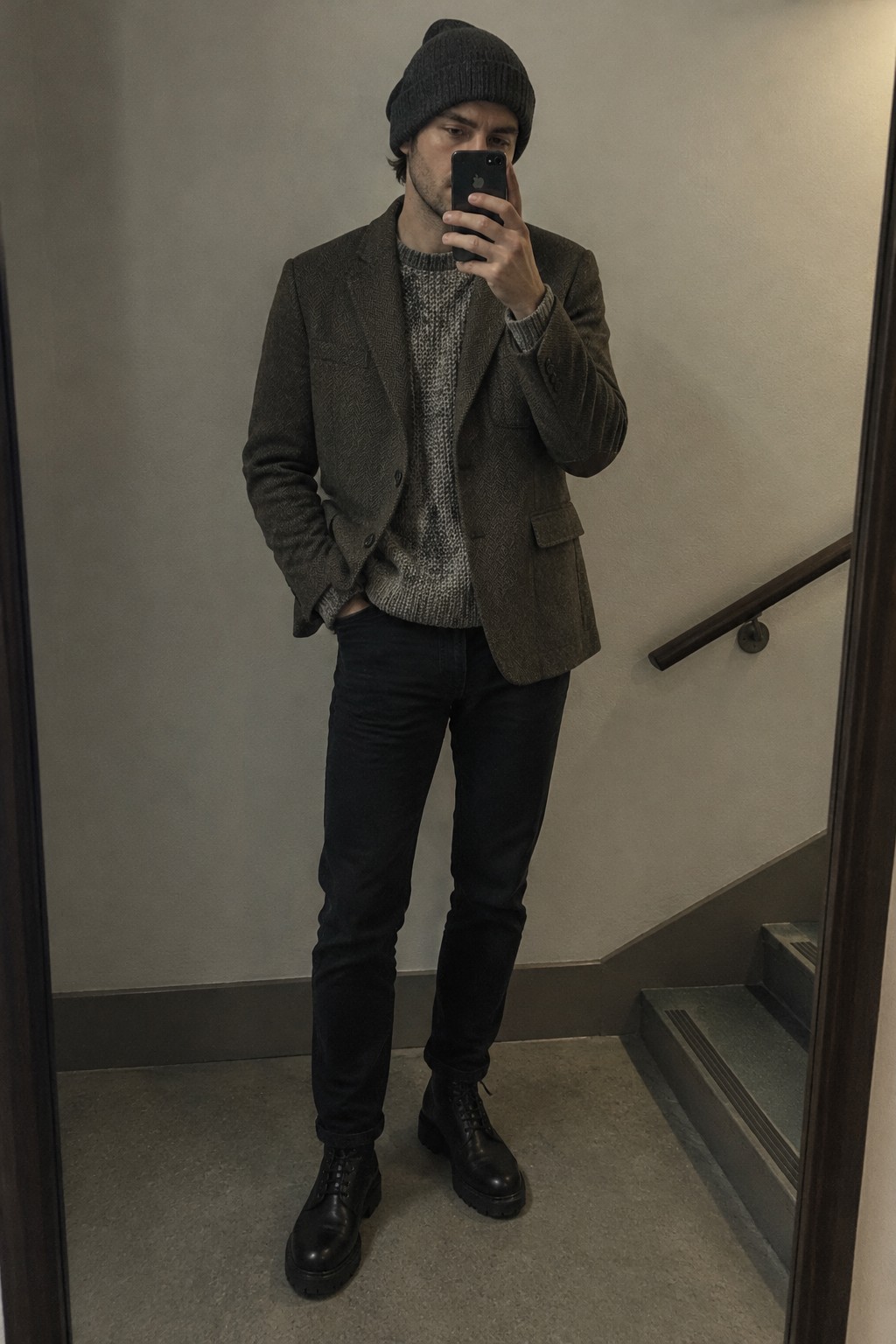 Man wearing a brown tweed blazer over a marled grey sweater with black jeans, a black beanie, and black lace-up boots.