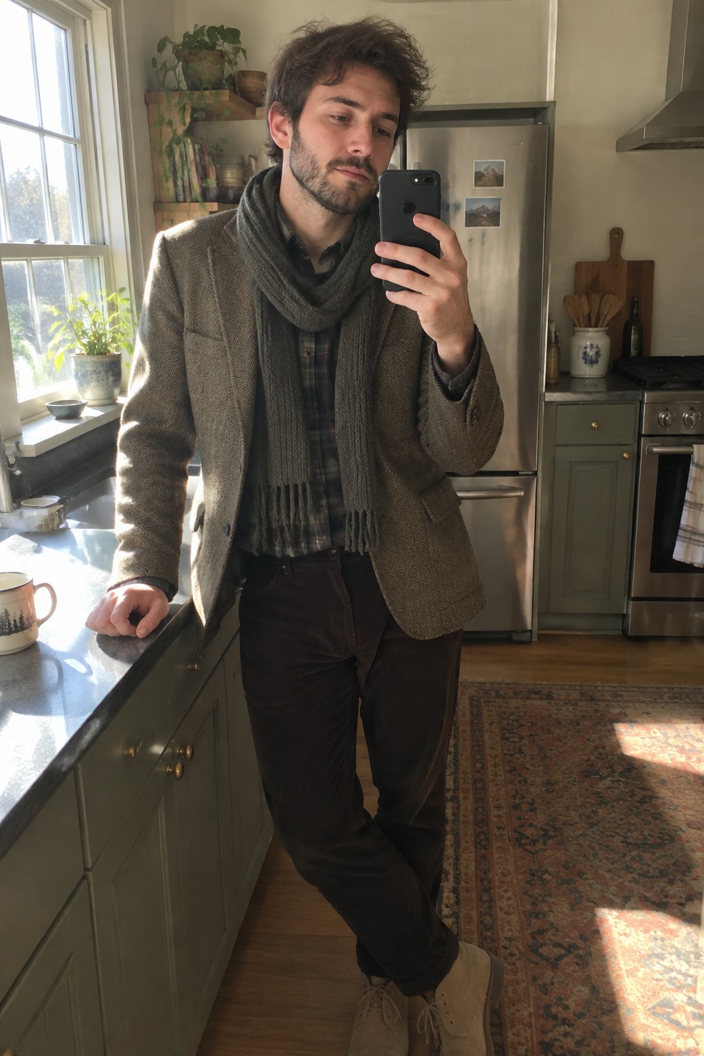 A man wearing a brown tweed blazer, checked shirt, long grey scarf, dark corduroy trousers and beige lace-up shoes.