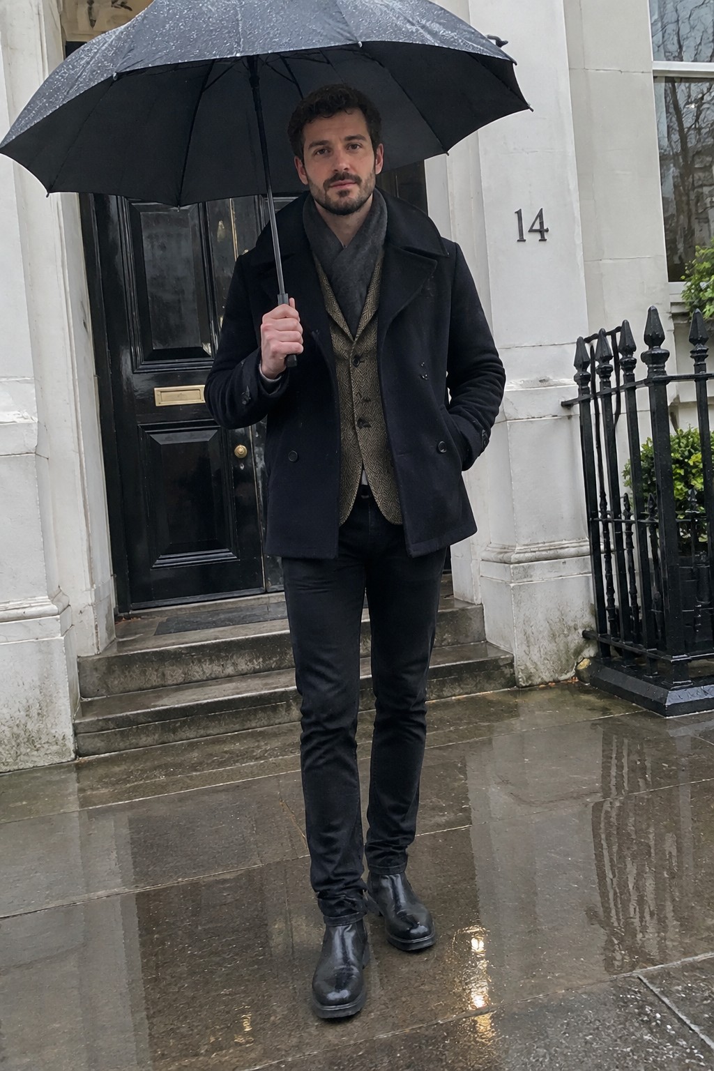 A man wears a brown tweed waistcoat, dark top, charcoal scarf, black wool coat, slim black trousers, and leather boots while holding a black umbrella.