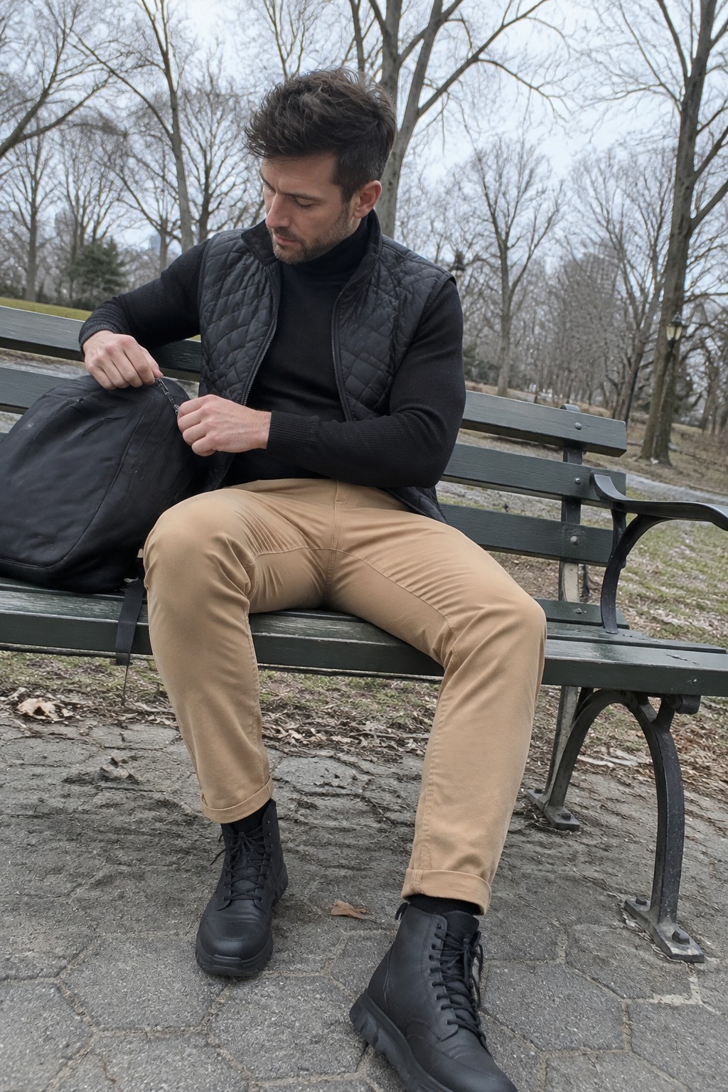 A man in a black turtleneck and quilted black vest, tan chinos, and black lace-up boots sits on a bench while opening a black bag.