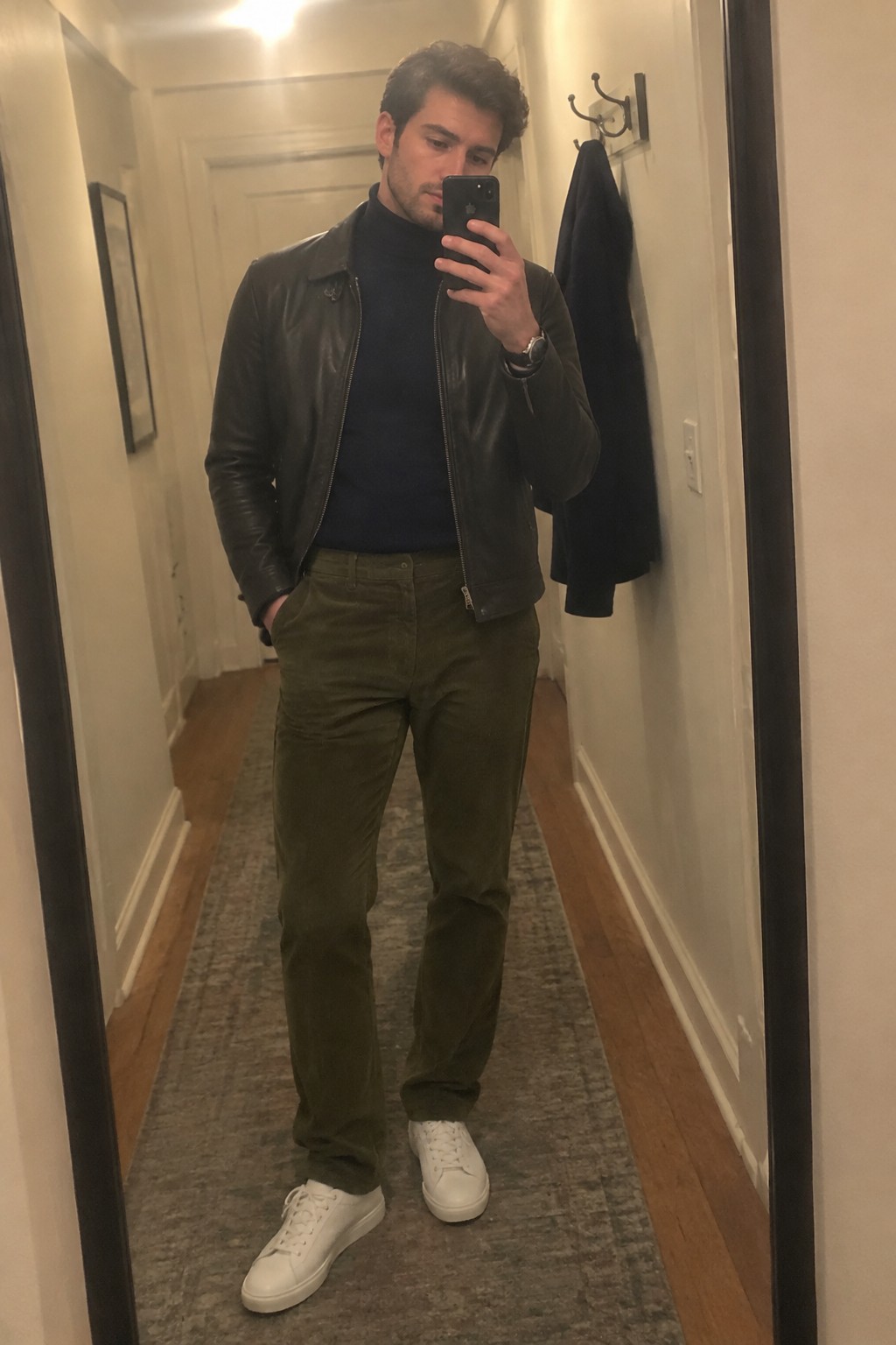 Man wearing a black turtleneck, dark leather jacket, olive corduroy trousers, white sneakers, and a wristwatch.