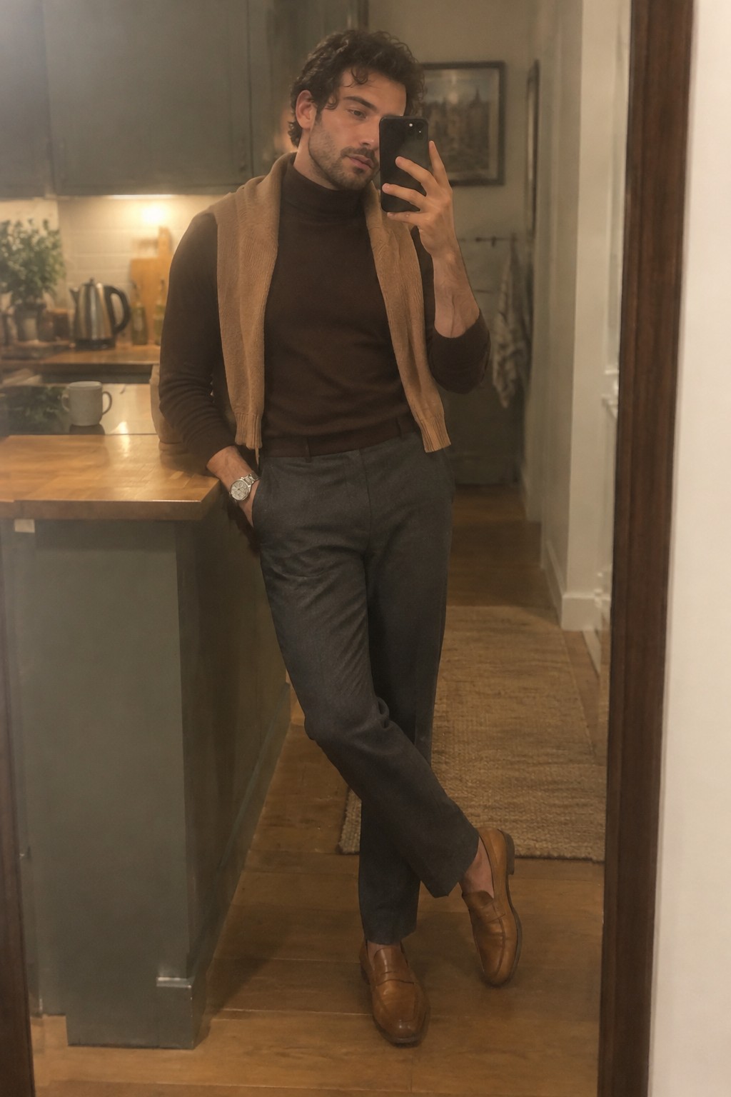 Man wearing a dark brown turtleneck, charcoal tailored trousers, a camel sweater draped over his shoulders, brown leather loafers, and a silver watch.