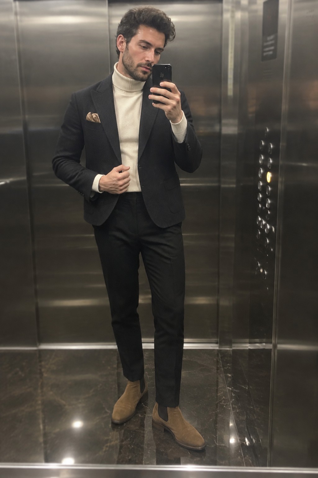 Man wearing a cream ribbed turtleneck, charcoal blazer and trousers, patterned pocket square, and tan suede Chelsea boots.